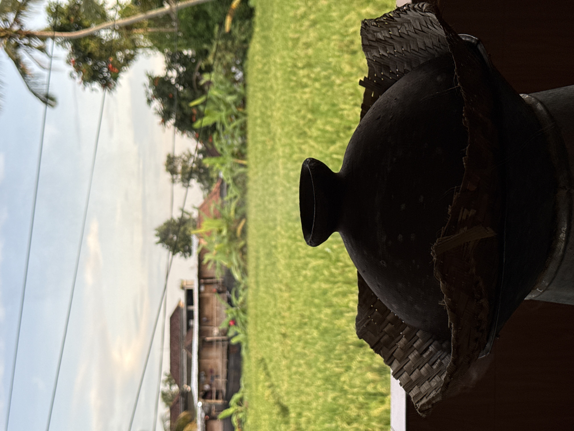 The image displays a serene rural landscape, likely in the Ubud District of Indonesia, presented with a unique perspective. In the foreground, positioned prominently on the right, is a dark, traditional pottery vessel, possibly an earthenware jar or pot. It's rendered almost entirely in silhouette due to backlighting, showcasing its rounded body and narrow neck. A rustic, woven material, resembling dried palm leaves or bamboo, partially covers or adorns the vessel, adding a textured, artisanal detail.
Stretching across the midground to the left is a vibrant expanse of lush green rice paddy, indicating an agricultural setting typical of the region. Beyond the rice field, in the background, are scattered tropical trees and foliage, along with glimpses of modest, rustic buildings, suggesting a village or small settlement. The sky above is a soft, hazy blue with lighter, cloud-like areas, suggesting either early morning or late afternoon light under overcast or partly cloudy conditions. Some power lines or wires are visible in the upper portion of the frame.
No specific activity is taking place; the image captures a tranquil moment in a natural setting. The overall mood is peaceful and evocative of traditional Indonesian rural life. There is no visible text in the image.