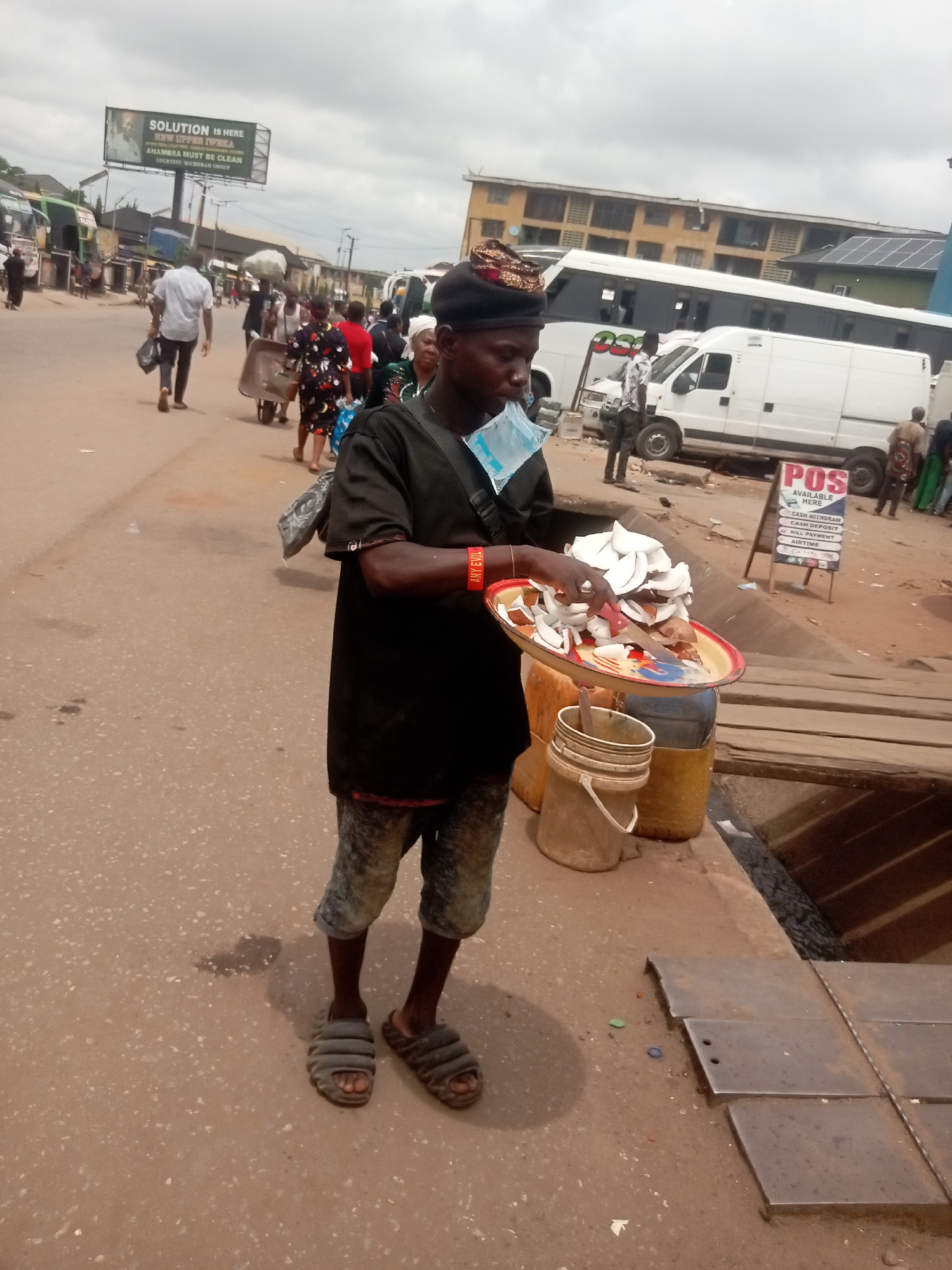 A medium shot depicts a man standing on the street in Onitsha, Nigeria, likely engaged in vending sliced coconut. The man, attired in a black t-shirt, patterned cap, and cutoff denim shorts, holds a tray piled with coconut slices in his left hand. A disposable plastic bag dangles from his neck. He is wearing slip-on sandals.
In the background, a bustling street scene unfolds, punctuated by buses, buildings, and pedestrians. A sign for POS (Point of Sale) services is visible, indicating a business nearby. An overhead billboard promotes the cleanliness of Anambra state, suggesting a civic initiative.
The setting is urban, with the infrastructure and commercial activity indicative of Onitsha's energetic street life. The presence of a water drain with loose metal plates near the vendor suggests infrastructure challenges.