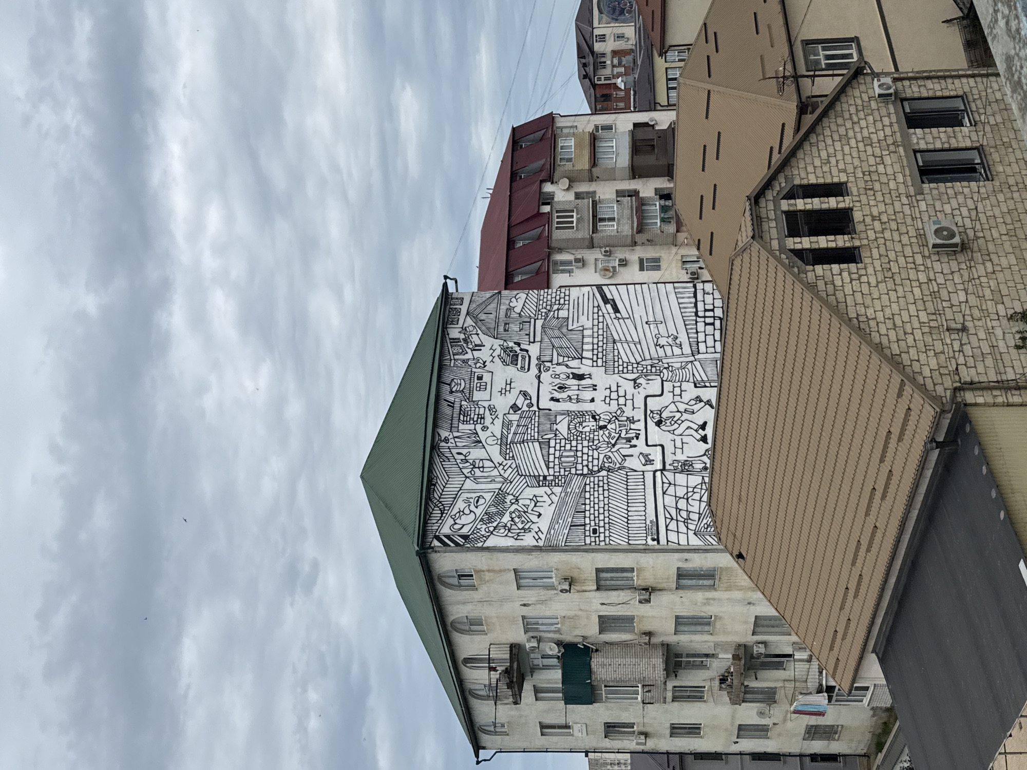 The image displays a cityscape with a focus on apartment buildings under a cloudy sky. The buildings are varied in architecture and age, with some featuring older, stucco facades and others having more modern brickwork or corrugated metal roofing. A striking feature is a large black and white mural covering a significant portion of one building's facade. The mural depicts a chaotic, busy scene with numerous small figures engaged in various activities, as well as representations of buildings and everyday objects.
The perspective is from a slightly elevated viewpoint, looking up at the buildings and the sky. The sky is overcast with soft, diffused light, suggesting it is either early morning or late afternoon, or simply a cloudy day. The overall mood is somewhat somber due to the weather, but the vibrant mural adds a touch of life and visual interest to the urban environment.
The image appears to be taken in Derbent, Russia, as indicated by the prompt. There is no discernible activity or event taking place in the immediate scene, but the mural itself suggests a narrative or commentary on urban life. Visible text is limited to a small signature or artist's mark on the mural itself, which is too small to be legible in the provided image. Other details include numerous air conditioning units mounted on the building walls and various windows, some with balconies.