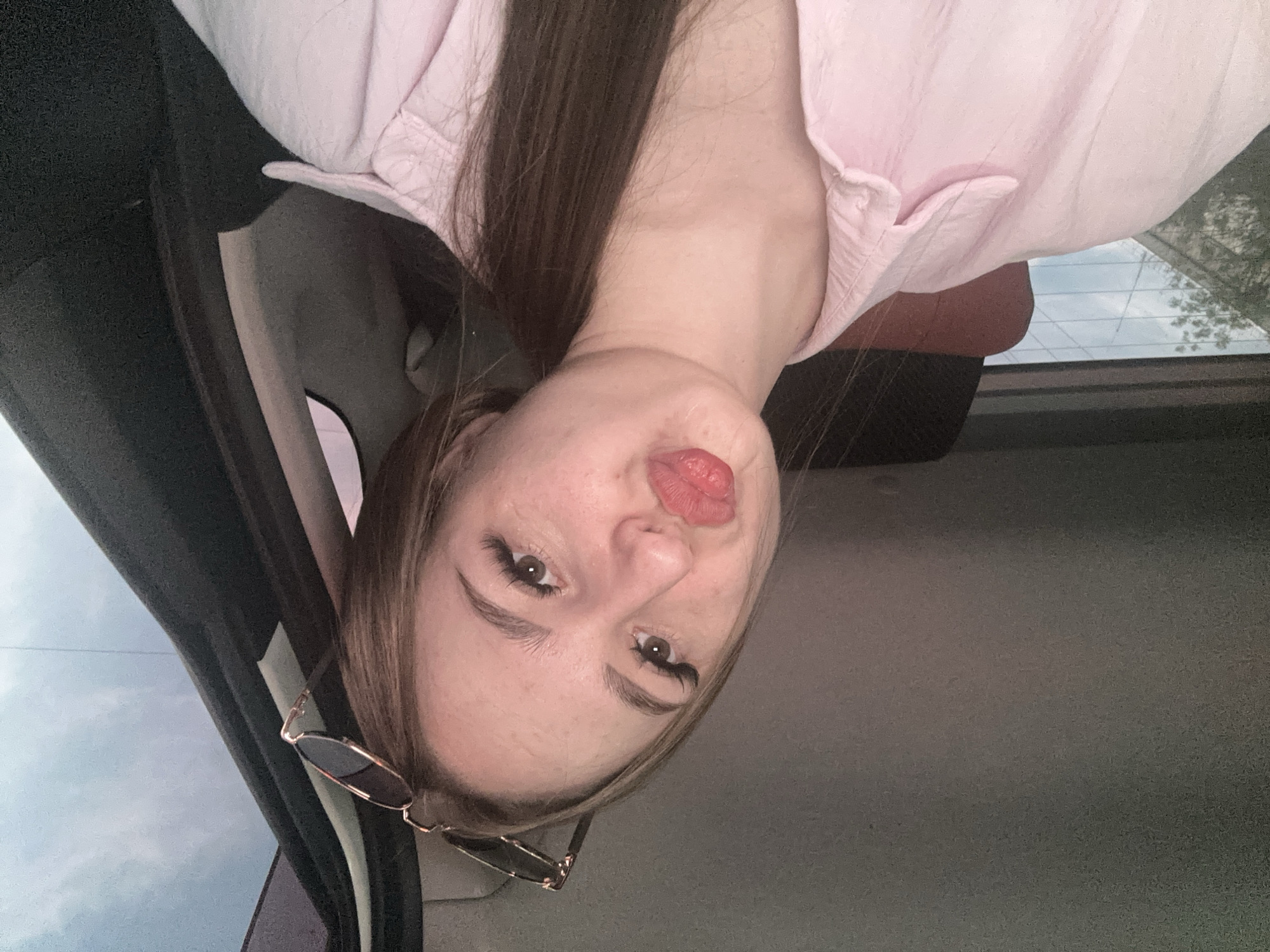 A young woman is the central subject of the image, captured from the chest up in what appears to be a selfie taken inside a car. She has long, straight dark brown hair, parted on her left side, which falls over her shoulders. She is wearing a light pink collared shirt. Her face is turned slightly towards the viewer, and she has a playful expression with her lips puckered and her tongue slightly extended. Her eyes are brown with prominent, dark eyelashes, and her fair skin shows some faint freckles. A pair of sunglasses with dark, round lenses and gold-toned frames is hooked onto the side of her head, near her right ear.
The setting is the interior of a vehicle, suggested by the dark car interior trim visible around her. Through the car windows, a cloudy or overcast sky is visible, indicating it is daytime. The back window, with its horizontal defroster lines, shows a reflection of tree branches against the sky. The overall impression is candid and casual. There is no visible text in the image.