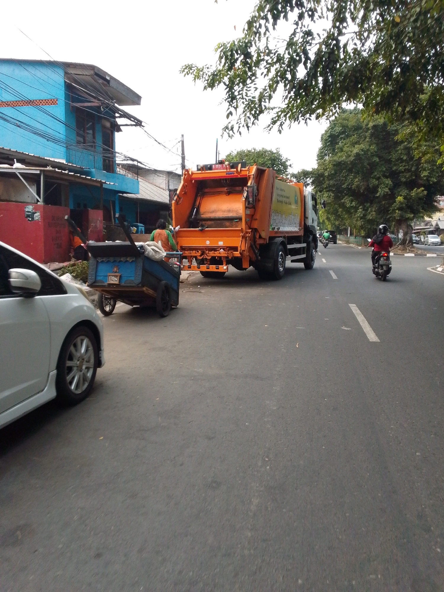 A street scene in East Jakarta, Indonesia, features an orange garbage truck on the right side of the road. The truck has a large compactor at the rear and is being driven on the asphalt road. In front of the truck, a person is seen pushing a blue handcart with black wheels, laden with items. To the left of the handcart, a white car is parked. In the background, a blue building with a tin roof and numerous overhead wires is visible, along with some trees and a distant view of other buildings. A woman on a scooter is riding in the same direction as the garbage truck on the right lane of the road. The overall environment appears to be urban, with evidence of residential buildings and commercial activity.