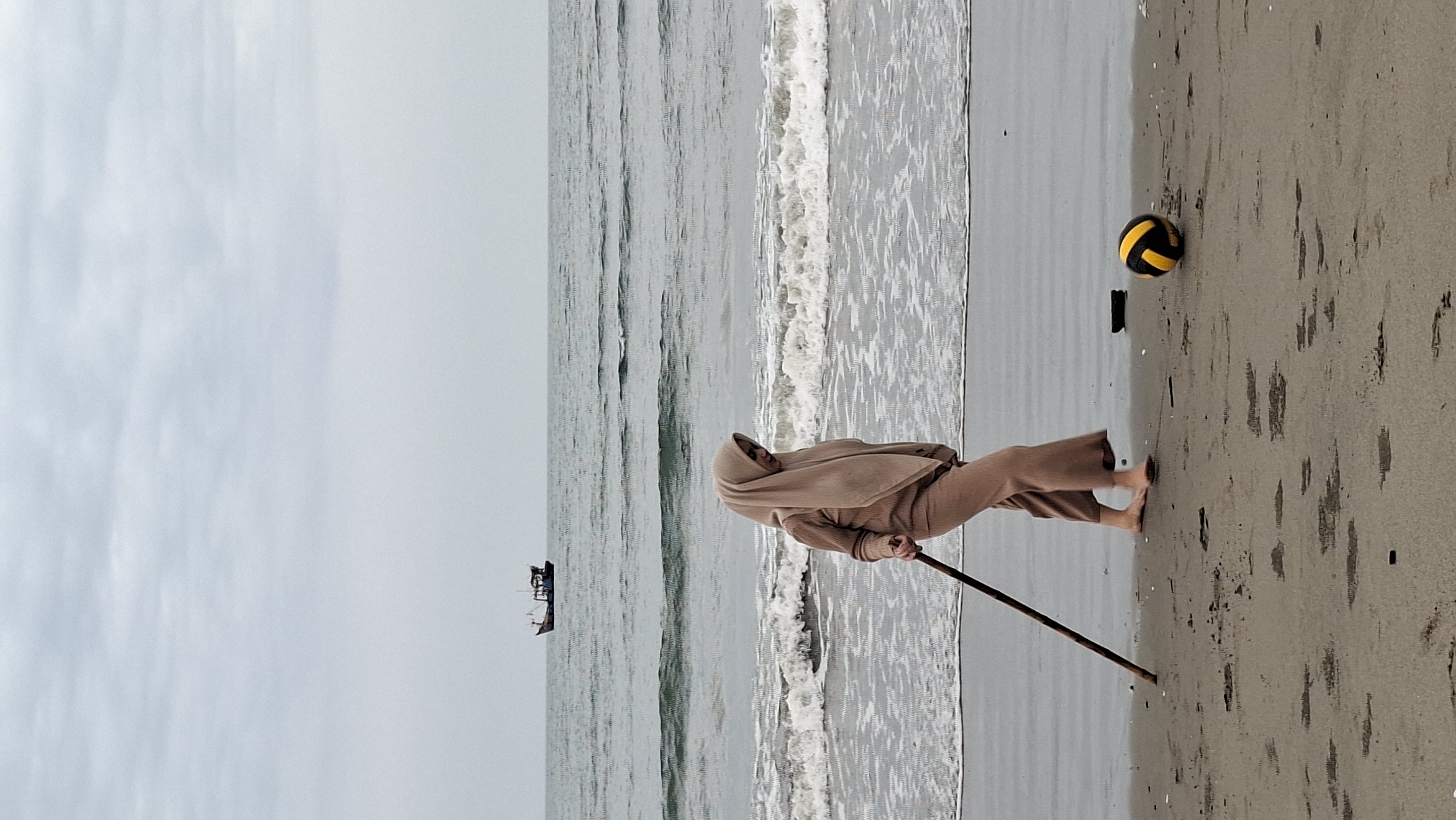 A woman wearing a beige hijab and matching loose-fitting clothing walks on a sandy beach in Lhokseumawe, Indonesia. She is holding a long wooden stick in her right hand, using it as a walking aid. Her left foot is bare and touches the wet sand near the water's edge. In the background, gentle waves roll onto the shore under an overcast sky. Further out at sea, a small fishing boat is visible on the horizon. To the right of the woman, a yellow and black volleyball lies on the sand. The beach appears to be sparsely populated, with no other people visible in the immediate vicinity. The overall atmosphere is calm and subdued, typical of a cloudy day at the coast.