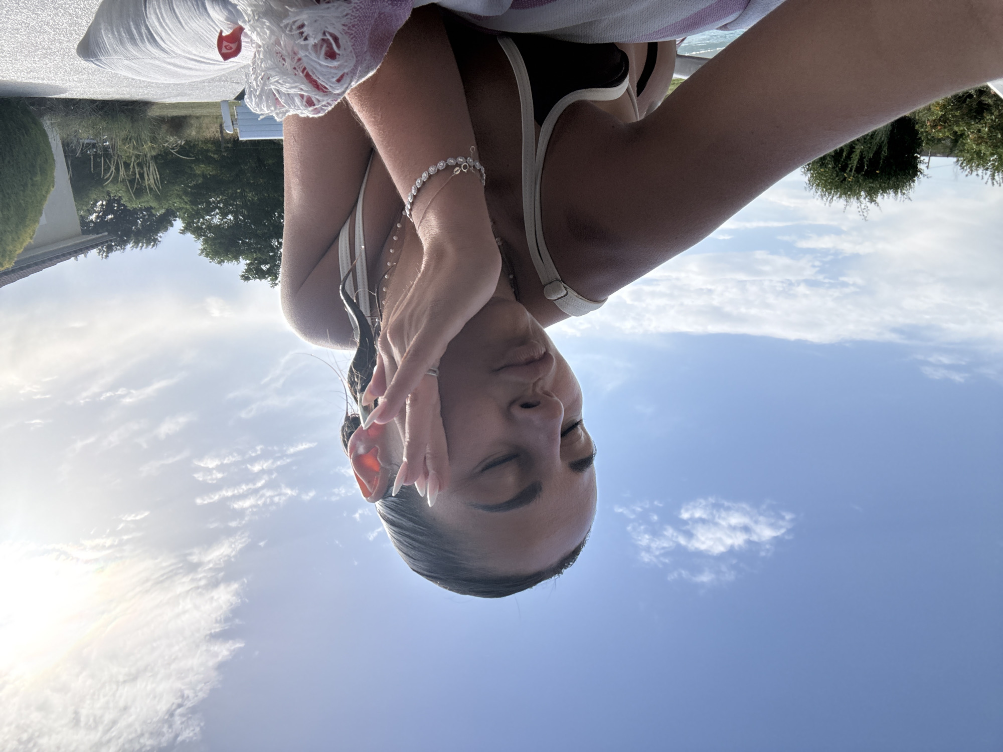 A young woman with wet hair is lying down outdoors, with her eyes closed and one hand touching her face. She is wearing a bikini and jewelry. The background shows a clear blue sky with some clouds, trees, and a house. The scene appears to be in Veauchette, France.