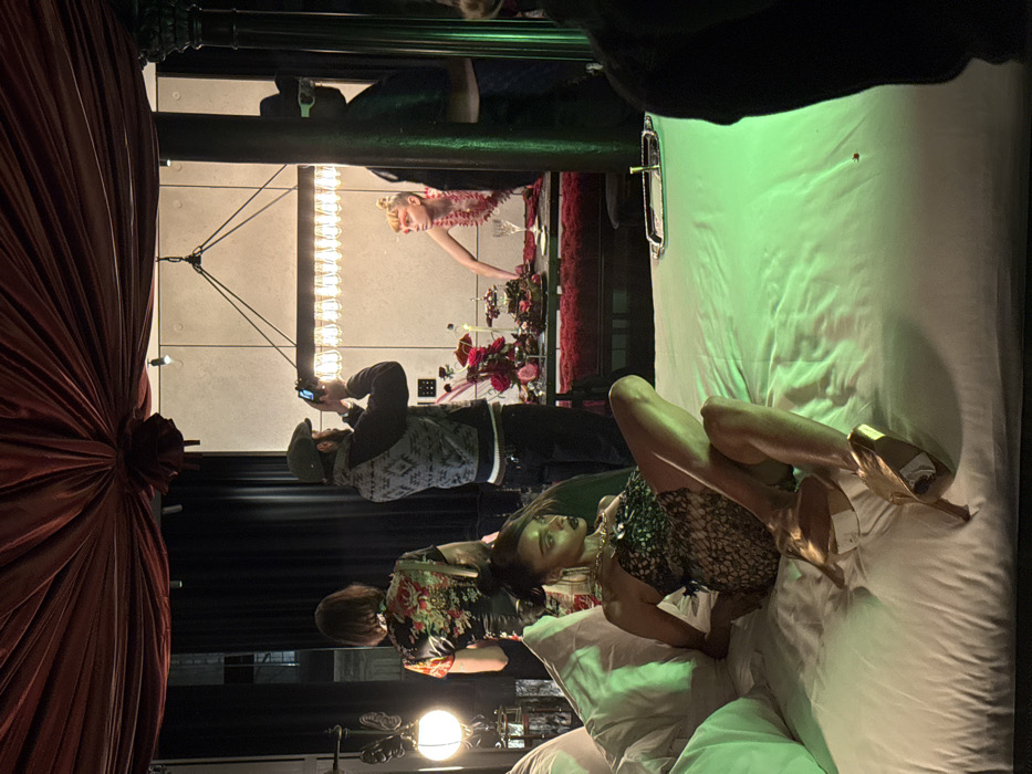 The image captures an indoor scene in the city of London, United Kingdom, depicting what appears to be a multi-person photoshoot or themed event. In the foreground, a dark-skinned female model lies on her left side on a large, white-linened bed, which is illuminated by a distinct green light. She wears a strapless, dark, patterned mini-dress or bodysuit, gold high-heeled shoes, large gold hoop earrings, and a gold necklace. Her dark hair is styled back with some strands framing her face, and she has dark lipstick. Her gaze is directed left, away from the camera. A silver tray with a slender metallic object rests on the bed to her right.
In the mid-ground, a bearded man wearing a dark flat cap, a grey patterned vest, and dark trousers holds a silver smartphone horizontally, aiming it towards the background to capture a photograph or video. Behind him, a woman with short, dark hair, seen from the back, wears a short-sleeved top featuring a vibrant red and green floral pattern on a dark background.
Further back, a light-skinned individual with blonde hair styled in a bun, wearing a red, spiky or textured top, leans over an elaborately decorated table. This table is covered in dark red fabric and adorned with multiple floral arrangements in deep red and pink hues, candles, small dessert or display stands, and wine glasses. Above the table hangs a rectangular chandelier with numerous exposed light bulbs, providing bright illumination. The background wall is a plain, light-colored, textured surface.
On the far right, partially visible, a man wearing a dark hat and distinctive white-rimmed sunglasses observes the scene. The ceiling above is draped with gathered dark red velvet fabric, creating a theatrical canopy. A decorative lamp with a butterfly motif and a spherical light is positioned on the far left, while a dark, ornate pillar stands on the far right.