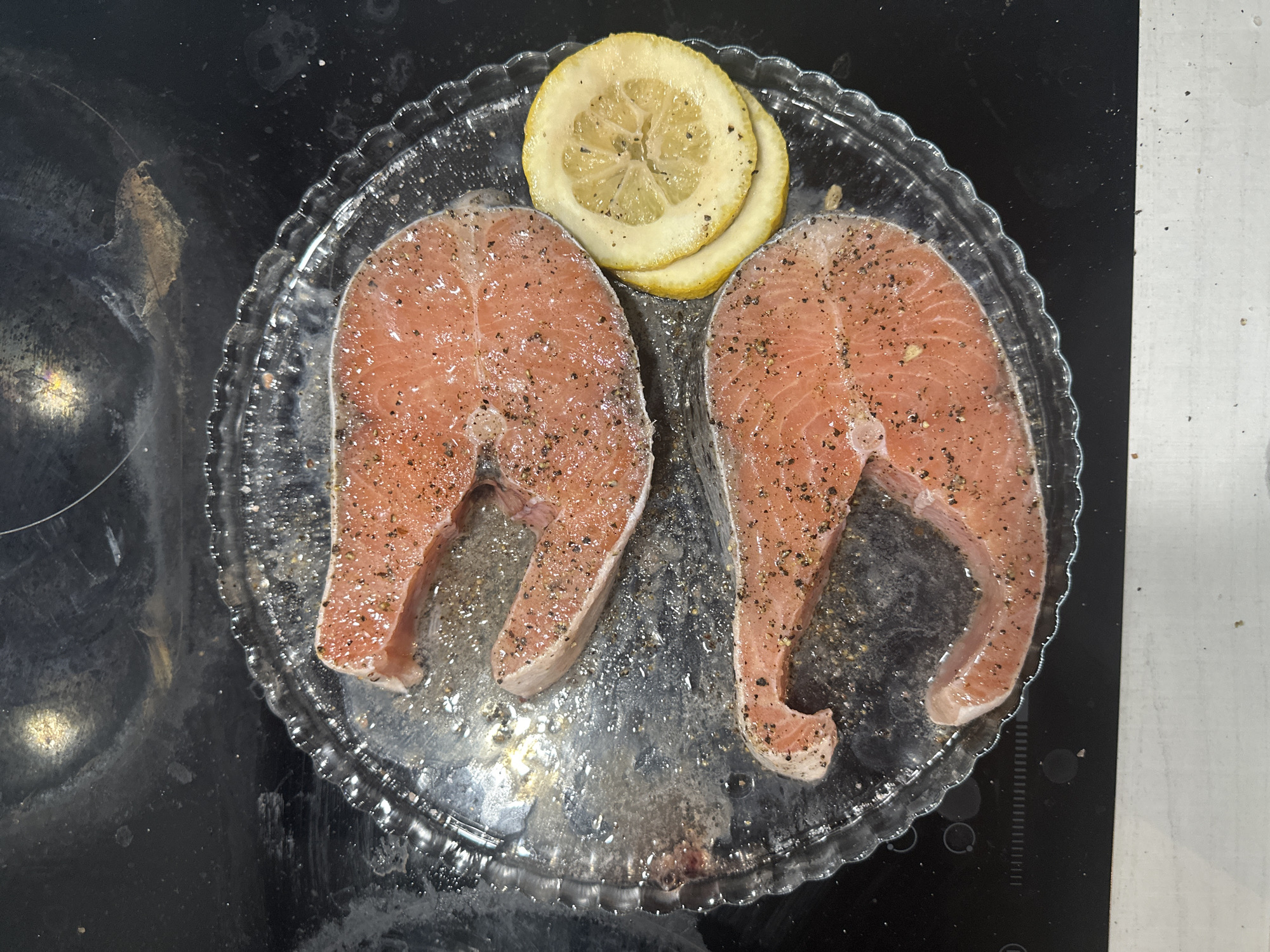 The image provides an overhead view of two raw salmon steaks, seasoned liberally with black pepper, placed on a clear, fluted-edge glass plate. Between and slightly above the salmon pieces are three slices of fresh lemon. The salmon is a vibrant orange-pink color, indicating its freshness. A thin layer of liquid, possibly oil or marinade, is visible on the plate beneath the fish.
The plate rests on a dark-colored stovetop, likely induction or ceramic, which shows some smudges, reflections, and remnants of previous use, such as circular outlines of burners and faint stains. To the right of the stovetop, a segment of a lighter-colored countertop or kitchen surface is visible. The scene captures a moment of food preparation in a kitchen setting. No people are visible, nor is any text present in the image. The location context is Khimki, Russia.