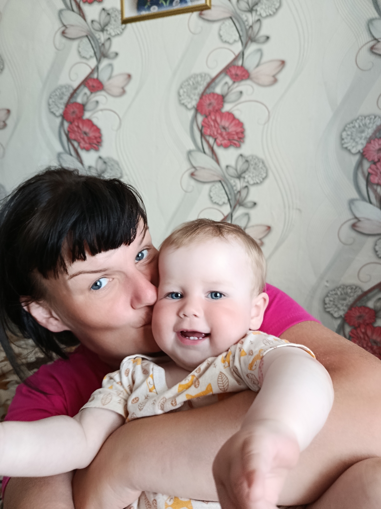 A woman with short dark hair and blue eyes is kissing a smiling baby with blue eyes and light brown hair. The woman is wearing a pink t-shirt and the baby is wearing a light-colored shirt with small orange animal designs. They are in front of a wall with a floral wallpaper pattern.
