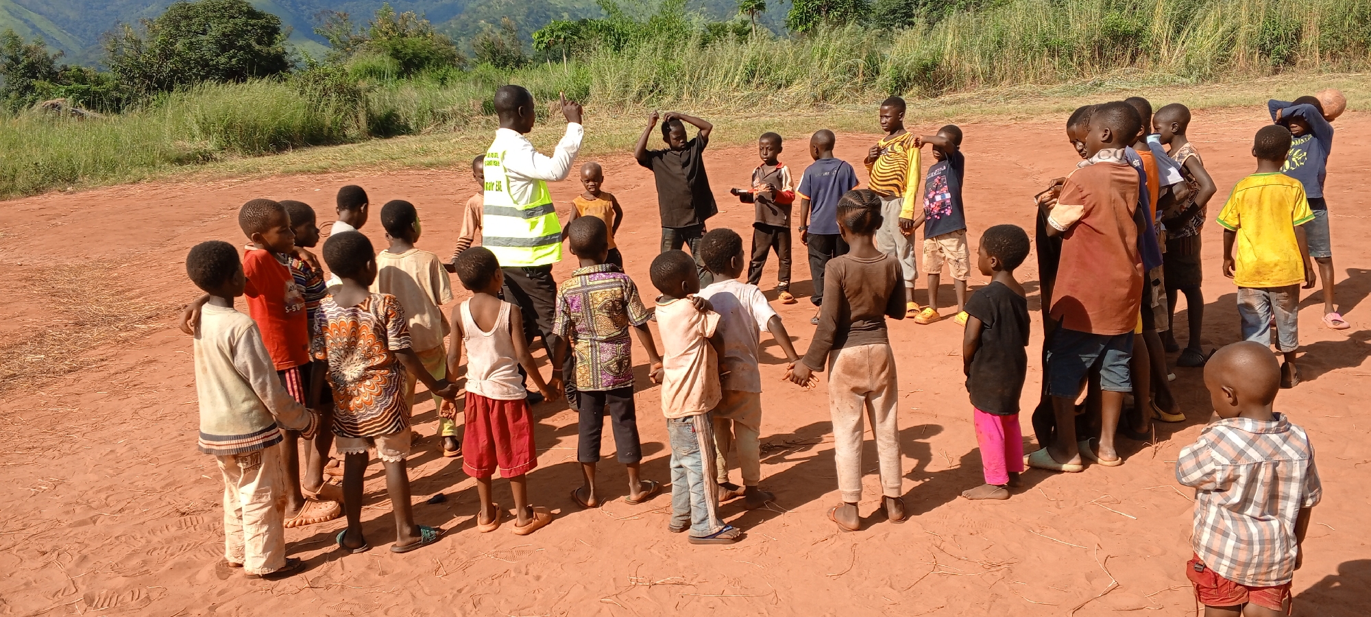 The image depicts a group of approximately 30 children gathered in a circle on a reddish-brown dirt field. An adult male, wearing a white shirt and a neon yellow safety vest, stands among the children with his right arm raised, seemingly instructing or leading the group. The children are dressed in a variety of casual clothing, including shirts, shorts, and pants in different colors and patterns. Some children are holding hands. The background shows a grassy area and a mountain range under a clear, sunny sky. The location is inferred to be near or in Ngalula, Democratic Republic of the Congo, based on the image context.