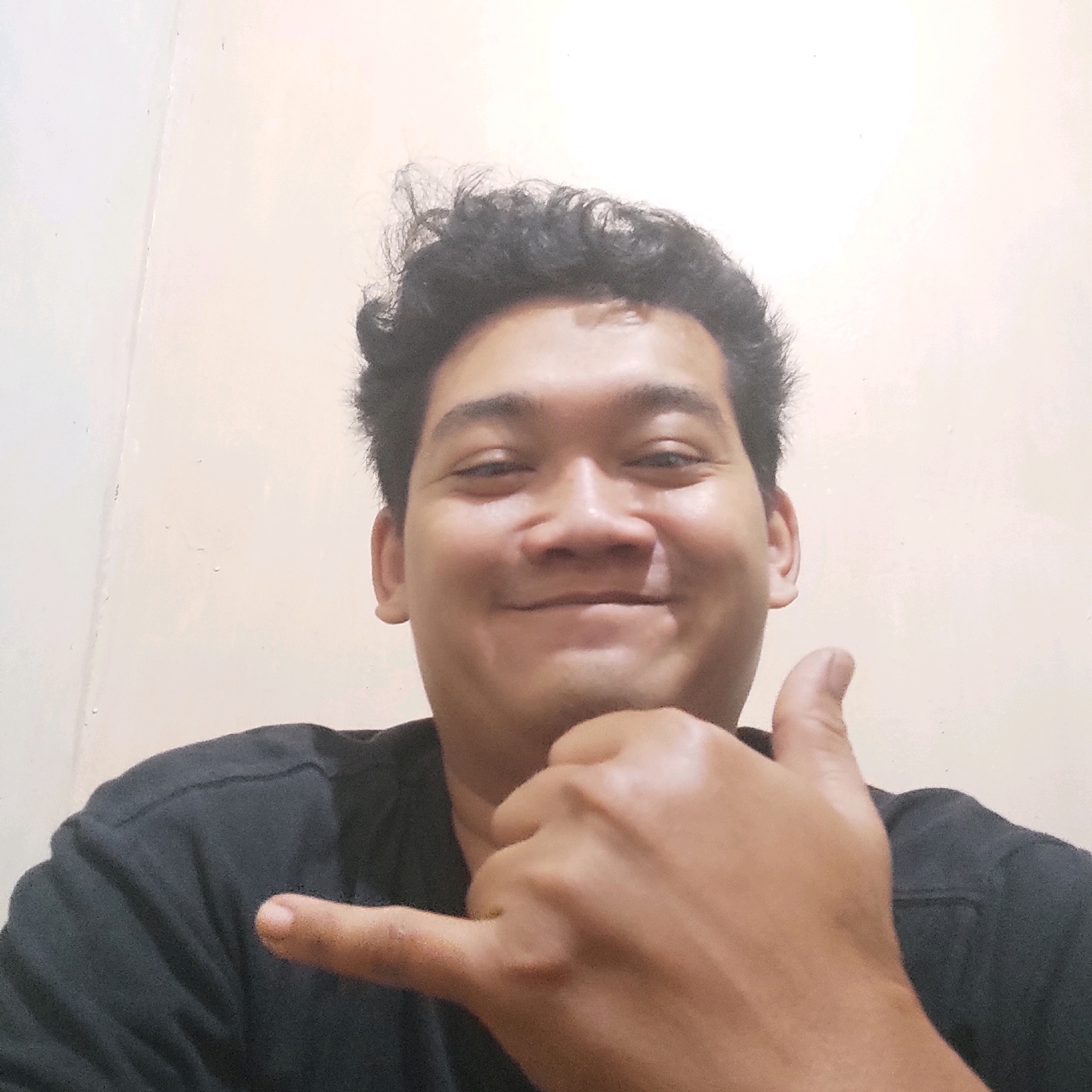A medium shot of a young man with dark, curly hair, wearing a black t-shirt, is captured from the chest up. He has a warm smile, with his mouth open slightly. His left hand is positioned in front of his chest, with his thumb extended upwards and his index finger curled towards his palm, forming a "shaka" or "hang loose" gesture. His right hand is held up near his face, with his index finger and thumb extended. The background is a plain, light-colored wall with subtle textured details. The lighting is soft and even, illuminating the man's face and hands.