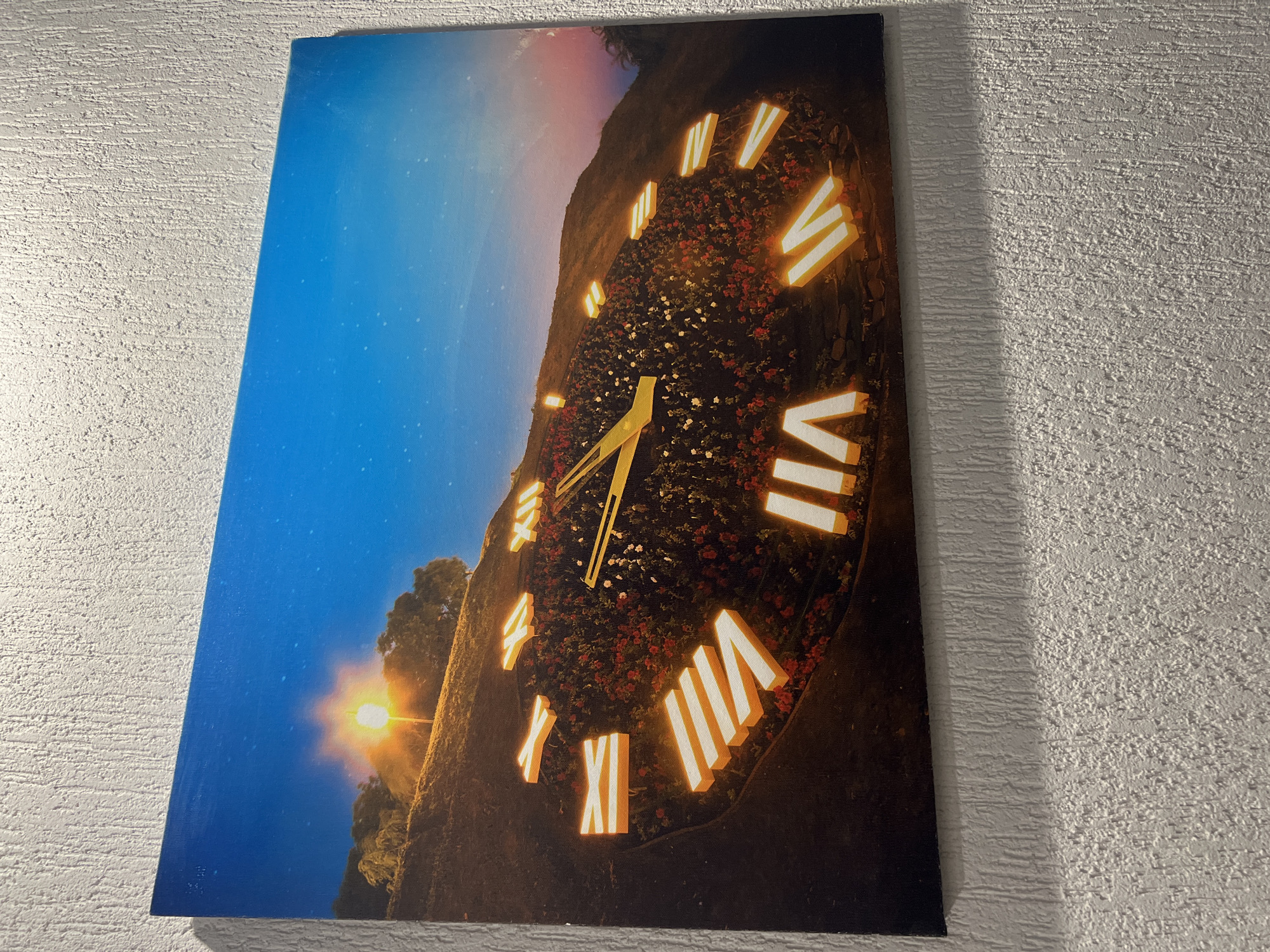 A canvas print depicts a clock face made of Roman numerals integrated into a flowerbed on a hillside. The clock face is illuminated, and the Roman numerals are outlined in a bright yellow light. The background shows a twilight sky with a bright light source on the left, possibly a streetlamp, and trees on the hillside. The print is hung on a textured white wall. The location in Yeysk, Russia is not discernible from the image.