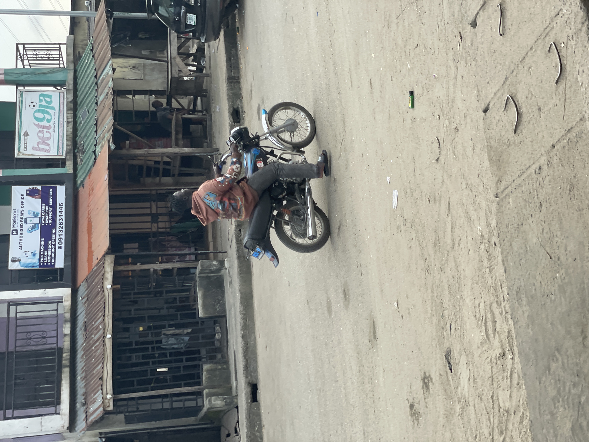 An individual rides a motorcycle on a dusty street in Ado, Nigeria. The person is wearing a brown hooded sweatshirt with a graphic on the back and dark pants. They are operating a blue and silver motorcycle. In the background, there is a row of buildings with corrugated metal roofs. One building has a sign that says "bet9ja" and another has a sign for "Moniepoint AUTHORISED BRM'S OFFICE". A black SUV is parked on the right side of the street. The ground is a mixture of dirt and some paved sections, with scattered debris.