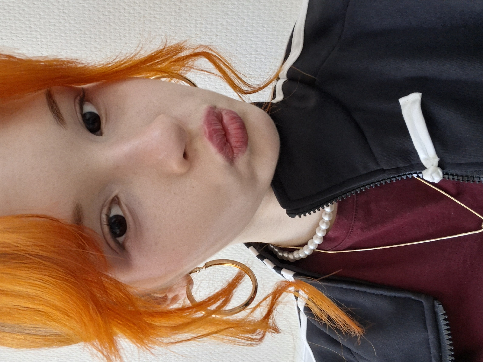 A close-up, tilted-angle shot depicts a person with vibrant orange hair. The individual has light skin, dark eyes, and pursed lips with red lipstick. They are wearing a black jacket with white stripes, layered over a maroon-colored top. Two necklaces are visible: a pearl necklace and a thin gold chain. A large gold hoop earring is visible in the left ear. The background appears to be a plain white surface. The provided context mentions the city of Chaikovsky, Russia, but there is no information in the image that confirms or denies this as the location.