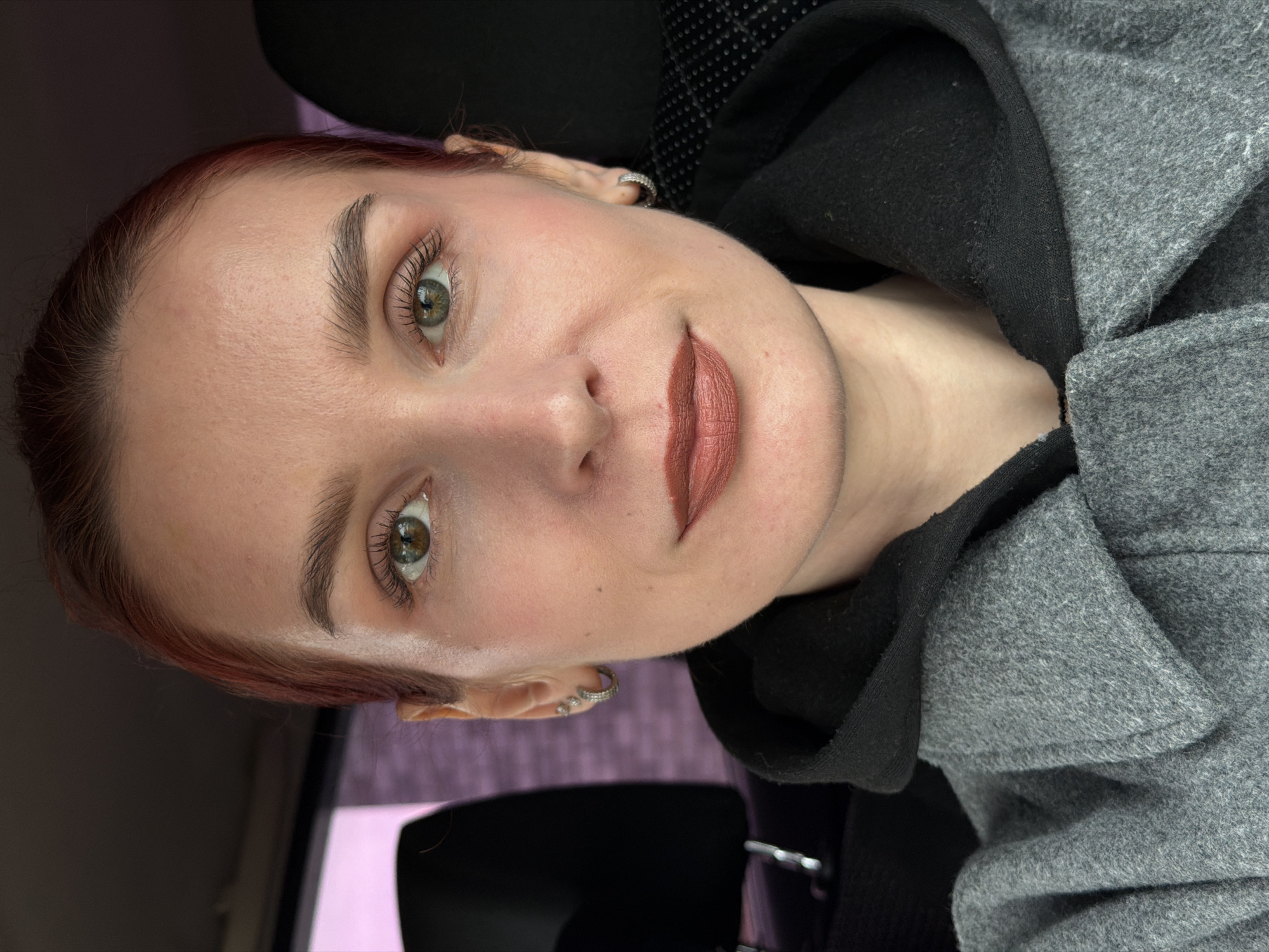 A portrait-style photograph shows a person with light skin, light green eyes, and reddish-brown hair pulled back. They are wearing makeup, including lipstick, and small earrings. The person is wearing a grey coat with a black hood. The background is a blurred combination of dark colors and muted pinks/purples, suggestive of an interior setting, possibly inside a car. There is no evidence suggesting a location in the city of Chaikovsky, Russia.
