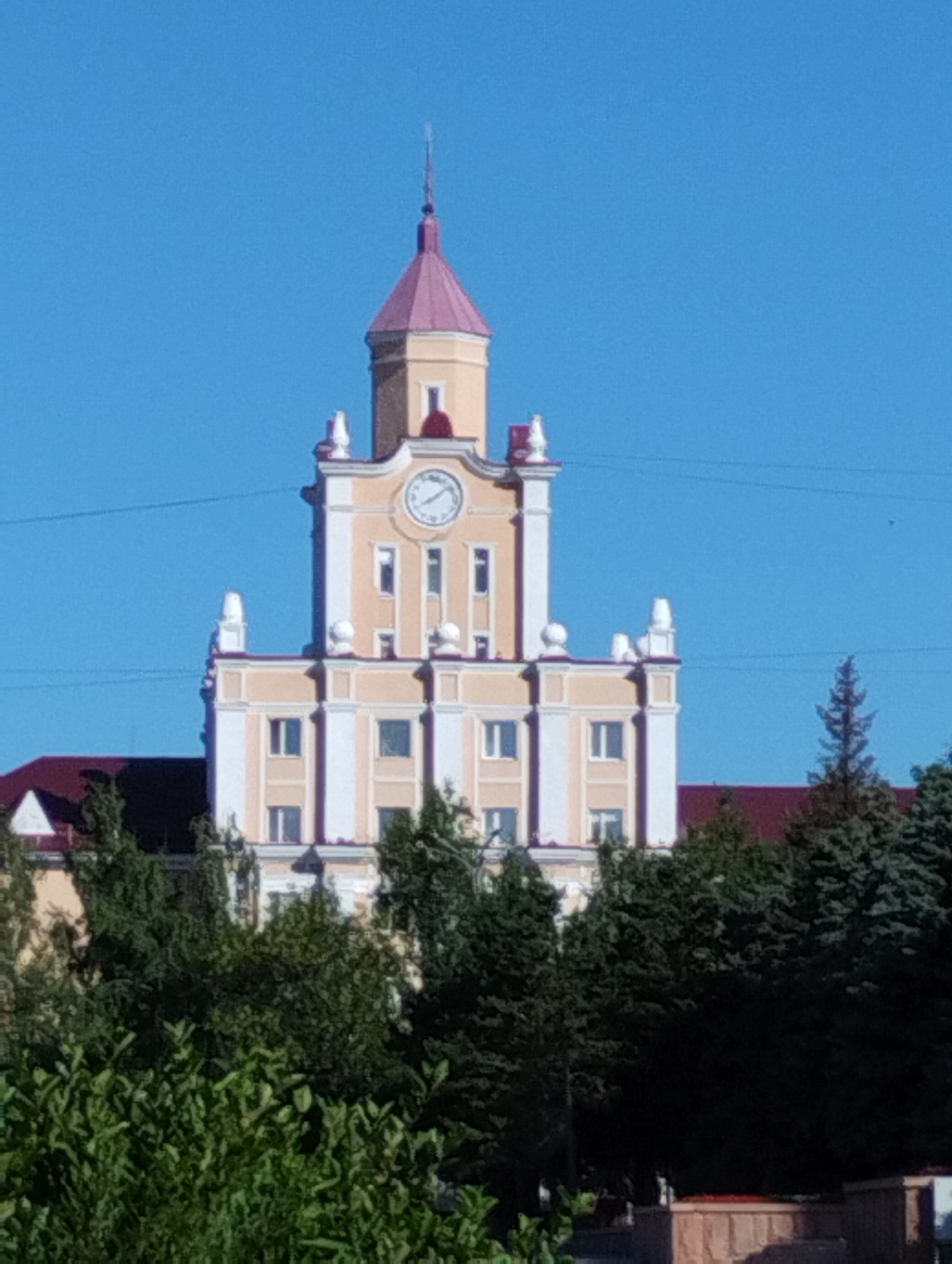 This is a medium-distance, eye-level, slightly blurry outdoor photograph of a tall, light-colored building with a pinkish-red pointed roof. The building has a tower in the center with a clock face. The clock face shows approximately 9:10. The building is adorned with white decorative elements and multiple rectangular windows. The sky is clear and blue. In the foreground and to the sides, there are dense green trees and bushes, obscuring the lower portion of the building. A dark red roof of another structure is visible behind the trees on the left side of the image. The building is likely a prominent architectural feature in the city of Kostanay, Kazakhstan.