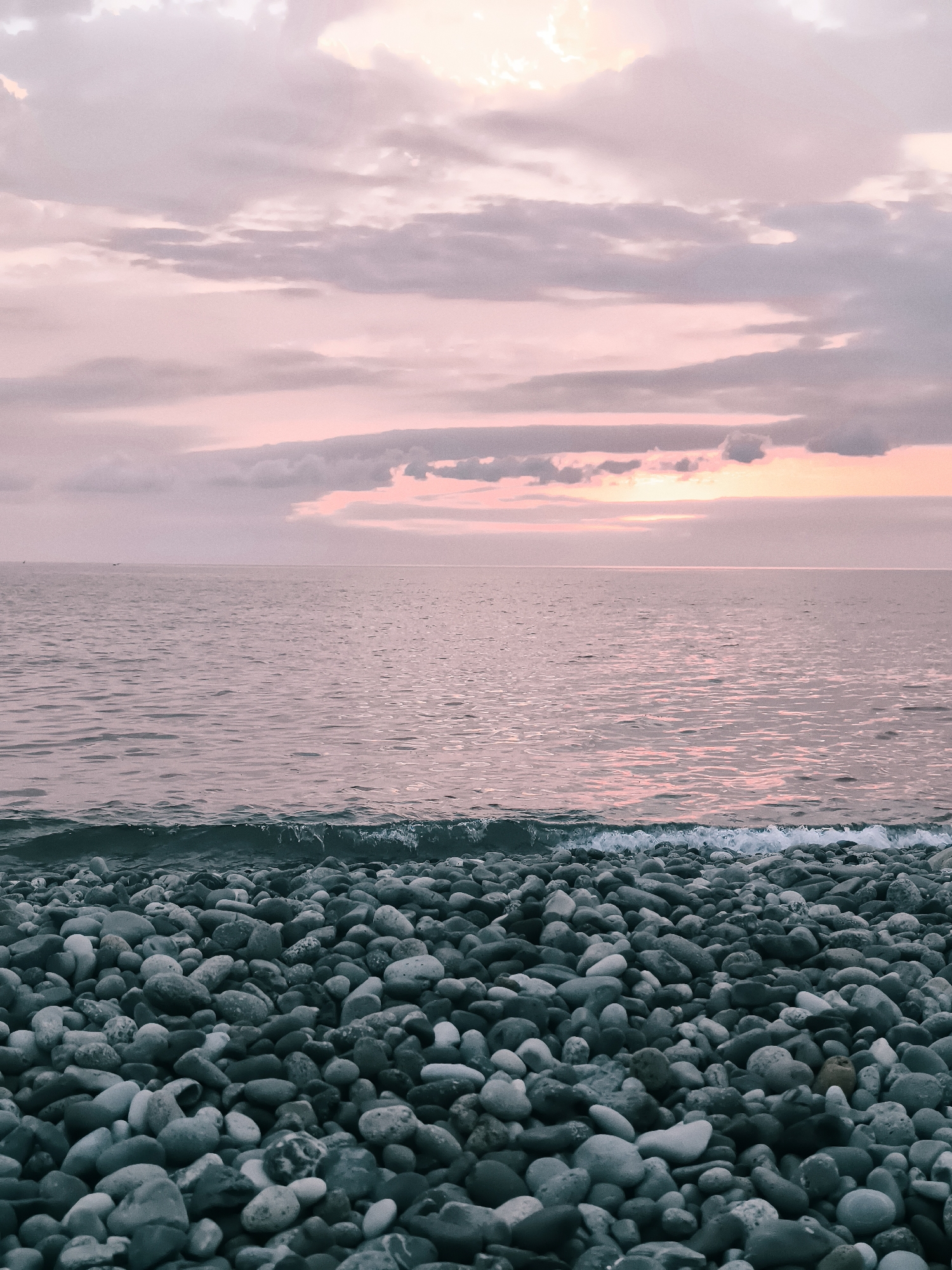 The image depicts a serene sunset scene at a pebble beach in Batumi, Georgia. The foreground is dominated by a dense collection of smooth, grey, and blue-toned pebbles of varying sizes, creating a textured and natural ground cover. A gentle wave, with white foam cresting, washes ashore at the bottom edge of the water, adding a dynamic element to the scene. The sea itself is a calm expanse of water, reflecting the soft, pastel hues of the sky. The horizon line is clear, separating the water from the sky. The sky is filled with a beautiful gradient of pinks and purples, with scattered clouds that are tinged with the warm colors of the setting sun. There are no people or man-made structures visible in the image. The overall atmosphere is tranquil and picturesque, characteristic of a peaceful evening by the Black Sea.