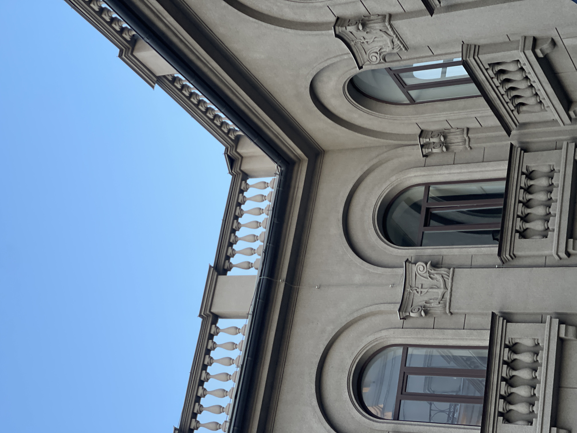 This media file presents an upward-looking view of a grand building facade against a clear, bright blue sky. The building, constructed from light grey stone or concrete, displays a classical or neoclassical architectural style with intricate detailing.
A prominent balcony with a balustrade, featuring decorative balusters, spans horizontally across the left side of the frame. Below this, the facade is punctuated by multiple arched windows, each framed in dark material, reflecting the sky and surrounding structures.
The building's surface is richly adorned with ornate architectural elements, including elaborate cornices, moldings, and carved decorations. Between and above the arched windows, detailed capitals or corbels with scrollwork and leaf motifs are visible. Smaller decorative balustrade elements also flank some of the windows. A dark drainpipe or similar conduit runs along a significant edge of the building structure.
The photograph is taken from a low angle, accentuating the building's height and architectural grandeur. The vivid blue sky and clear illumination indicate it is daytime. There are no people or specific activities depicted in the image, nor is any text visible. While the provided context suggests the location is Krasnoyarsk, Russia, the image itself does not contain explicit geographical cues.