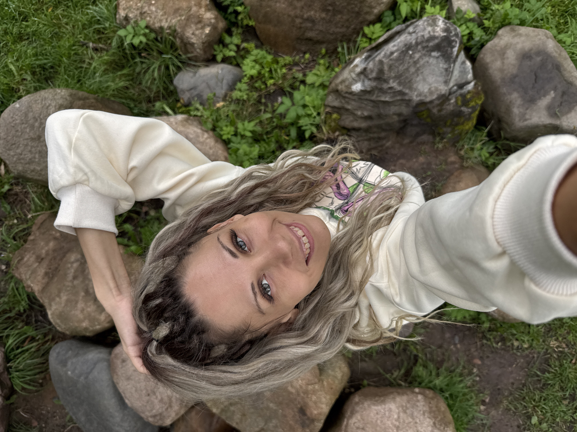The media file is a high-angle selfie of a young woman with long, wavy blonde hair. She is wearing a cream-colored sweatshirt and appears to be outdoors in a natural setting, surrounded by large rocks and green grass. Her head is tilted, and she is smiling at the camera, touching her hair with her left hand. Her eyes are blue, and she has on eye makeup. The background suggests a park or garden area. The lighting indicates it is daytime, and the weather appears to be mild, suitable for being outdoors. There is no visible text in the image. The location context is Rusanovo, Russia.