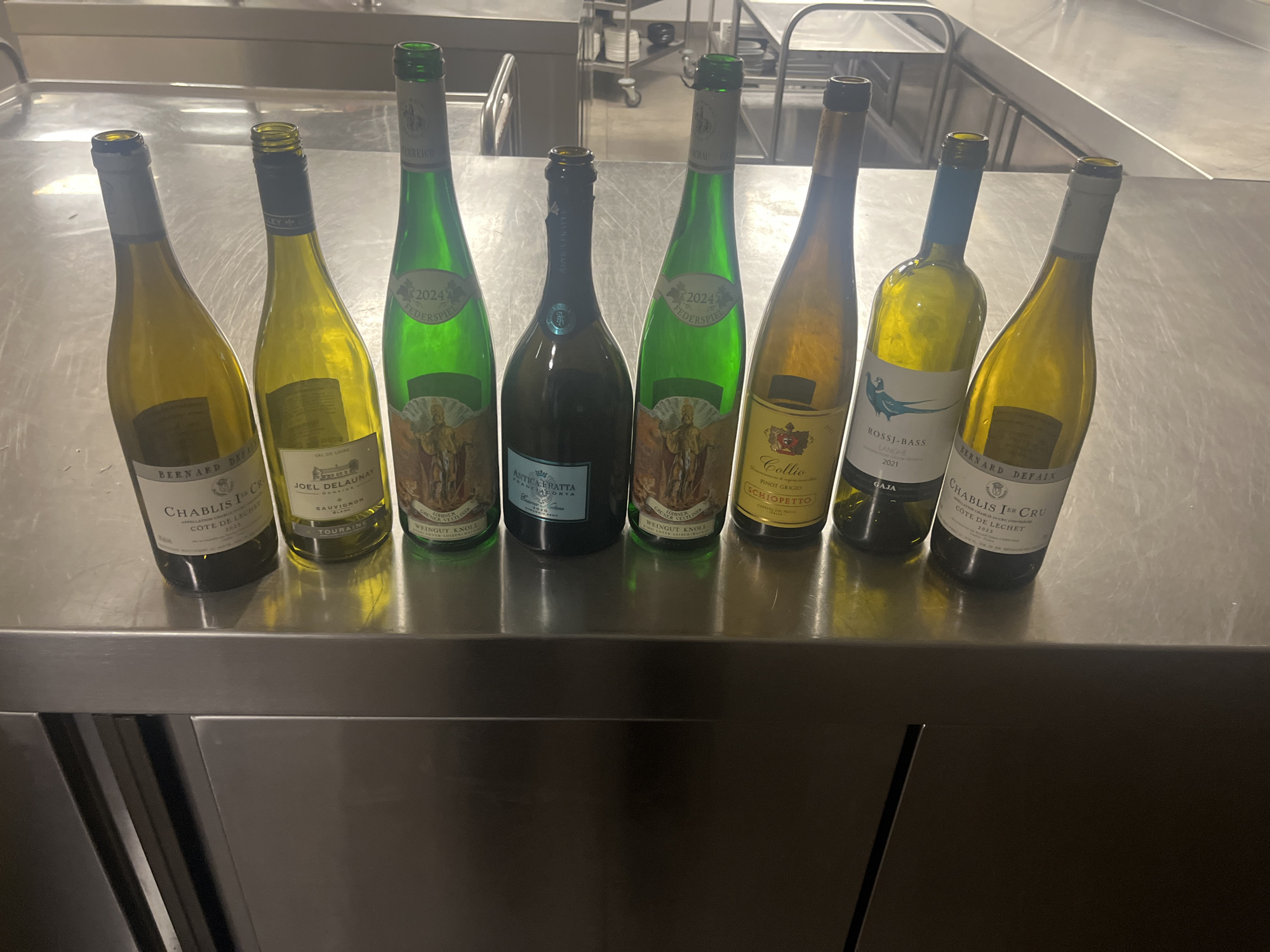 Eight bottles of predominantly white wine, including one sparkling wine, are displayed in a line on a highly reflective stainless steel countertop. The setting appears to be a professional kitchen or restaurant, characterized by extensive stainless steel surfaces, industrial-style shelving, and a utilitarian environment, possibly located in Sofia, Bulgaria. Artificial lighting illuminates the scene, casting reflections on the bottles and the metallic counter.
The wines, from left to right, include:
1. Bernard Defaix Chablis 1er Cru, Côte de Léchet 2023.
2. Joël Delaunay Sauvignon Blanc from Touraine, Val de Loire.
3. Weingut Knoll Grüner Veltliner Federspiel 2024 from Austria.
4. Antica Fratta Franciacorta (possibly 2020 vintage), in a darker bottle, indicative of sparkling wine.
5. A second bottle of Weingut Knoll Grüner Veltliner Federspiel 2024.
6. Schiopetto Collio Pinot Grigio.
7. Gaja Rossj-Bass Langhe 2021.
8. A second bottle of Bernard Defaix Chablis 1er Cru, Côte de Léchet 2023.
The bottles vary in glass color from clear to yellow-green, green, amber, and dark green. Visible text on the labels specifies the producer, appellation, grape variety, and vintage for most bottles. The arrangement suggests a wine selection for tasting or service.