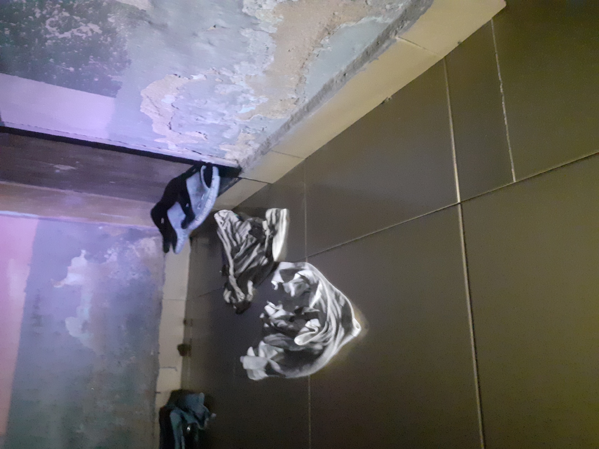 The image depicts a dimly lit interior space, likely a room, with tiled flooring. On the floor, there are several piles of crumpled clothing, appearing to be white and dark-colored garments. In the background, a pair of black and white flip-flops are visible, placed on top of some dark fabric. The walls are rough and weathered, with peeling paint, and a light purple hue is cast on the upper portion of the wall on the left. The overall environment suggests a state of disarray or abandonment. It is not possible to determine the specific location within Jalingo, Nigeria, from the visual information provided.
