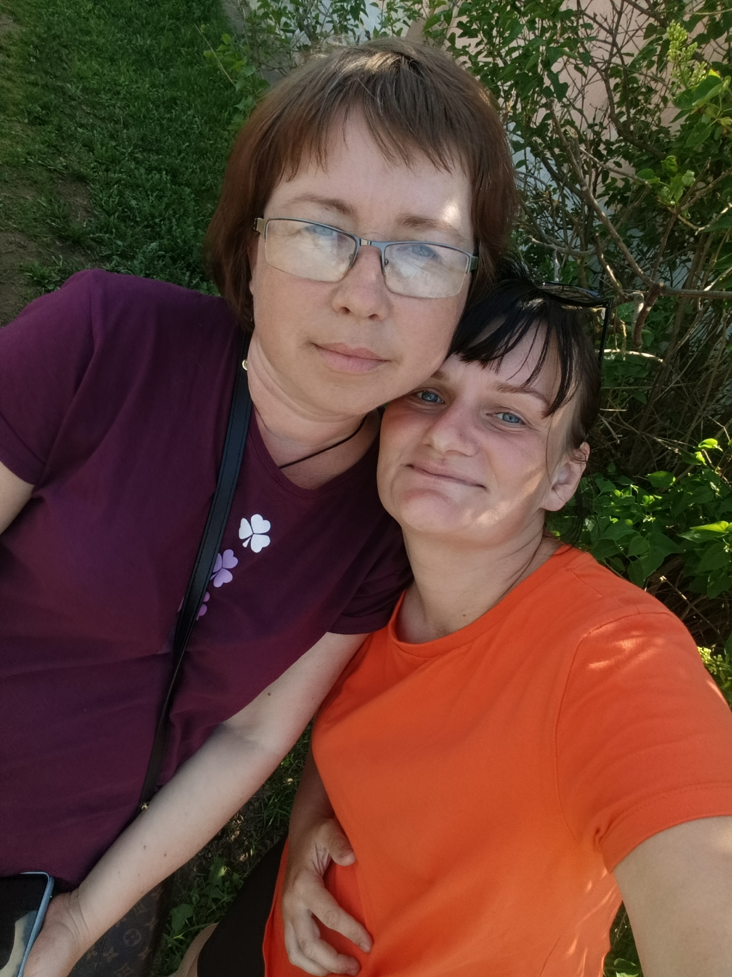 Two women are posing for a selfie outdoors. The woman on the left wears glasses and a maroon t-shirt with shamrock-like designs. The woman on the right wears an orange t-shirt. Green foliage is visible in the background. Based on the prompt, this image was taken in the city of Фоки, Russia.
