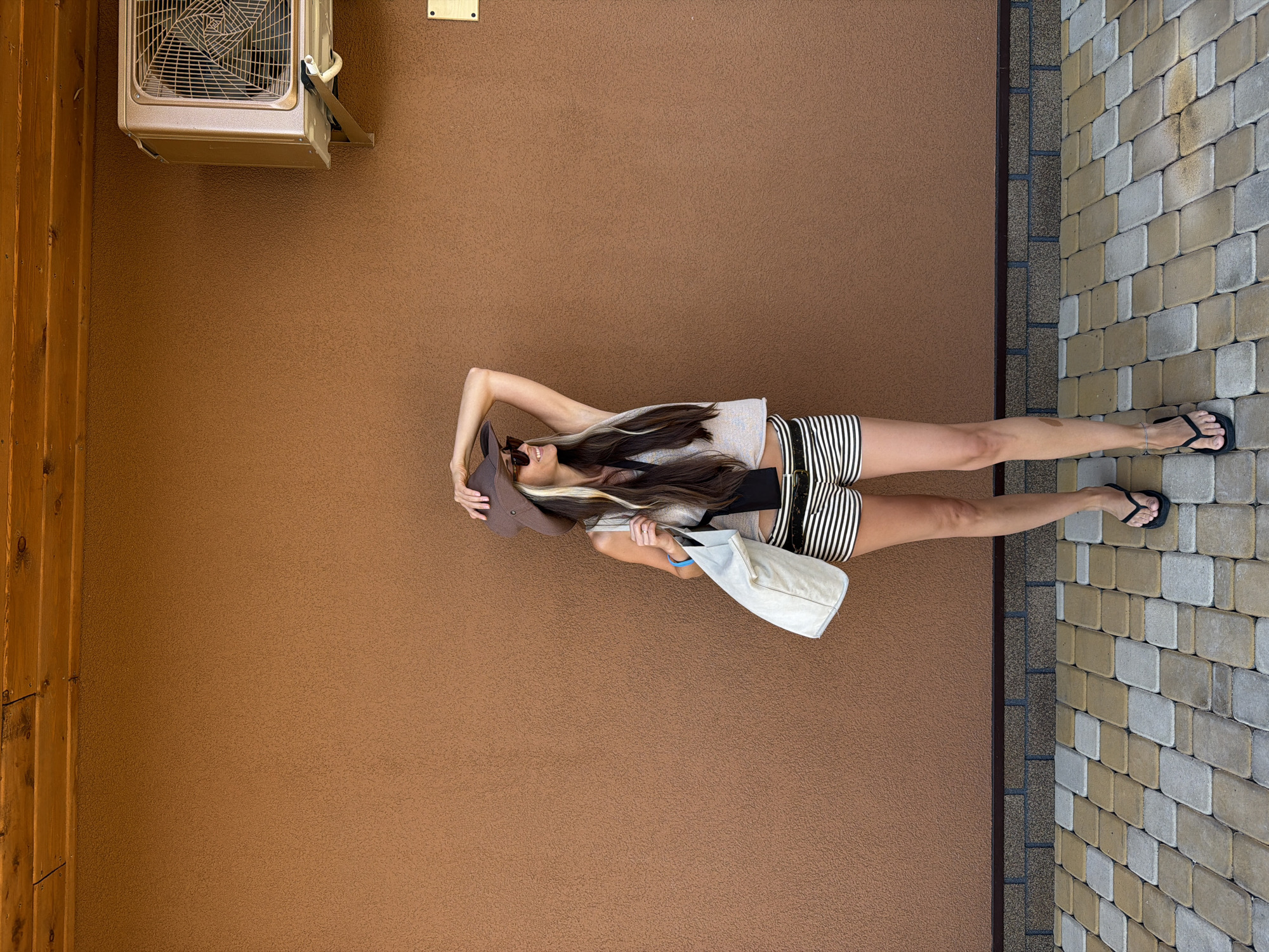 A young woman stands outside against a brown textured wall. She is wearing a wide-brimmed hat, sunglasses, a sleeveless top, and striped shorts. She carries a light-colored tote bag over her shoulder and wears flip-flops. Her left hand is raised, resting on her hat. To the right of the woman, mounted on the wall, is an air conditioning unit. The ground in front of the wall is paved with rectangular stones in alternating shades of tan and gray. The scene appears to be outdoors, possibly in a courtyard or along the side of a building. There is no identifiable information in the image to link it to Kamyshin, Russia.