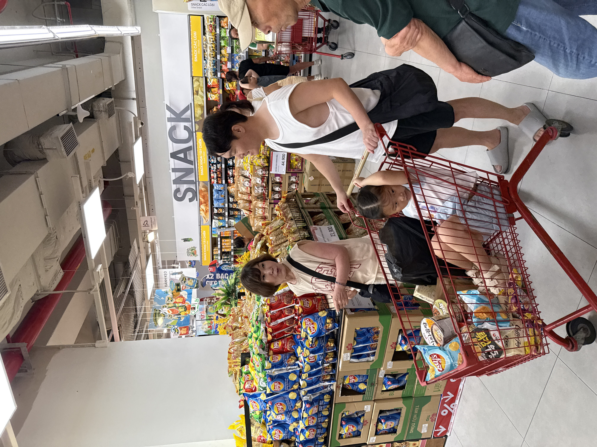 In a brightly lit supermarket aisle in Nha Trang, Vietnam, a family is seen shopping for snacks. A man in a white tank top pushes a red shopping cart filled with various snack bags and a cup of noodles. A young girl sits in the cart, looking down. To the left, a woman in a light-colored t-shirt stands near a display of Lay's potato chips. The aisle is lined with shelves stocked with a wide assortment of bagged snacks, with signs indicating "SNACK" and categories like "SNACK KHOAI TAY CHIEN" (Fried Potato Snacks). In the background, other shoppers are visible, and the overall environment suggests a busy retail setting.