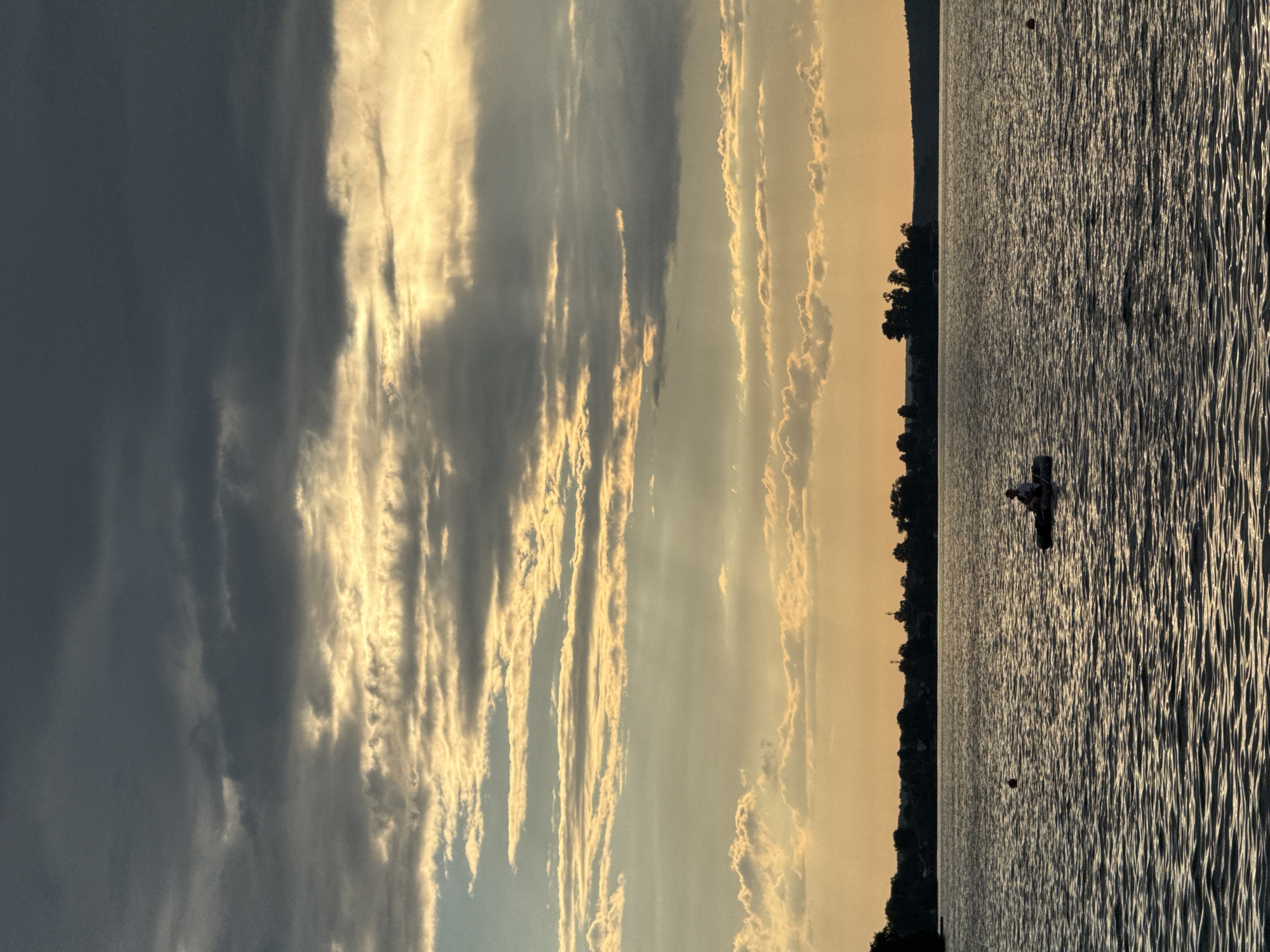 The image depicts a serene lake scene, likely Votkinsk Reservoir in Votkinsk, Russia, during the late afternoon or early evening. The sky is dramatic, with a mix of dark, brooding clouds in the upper half and streaks of golden sunlight breaking through in the middle and lower sections, illuminating the layered cloud formations. The sun's rays create a diffused, ethereal glow. Below the sky, a vast expanse of water stretches across the frame, rippling with gentle waves that catch the light. In the distance, a silhouetted tree line marks the shore, with hints of buildings and possibly a radio tower visible against the horizon. The water's surface is dark, reflecting the muted colors of the sky. A lone figure is seated in a small inflatable boat or raft in the middle ground, facing towards the right side of the image. Another small, round, orange buoy is visible to the left of the boat. The overall atmosphere is peaceful and slightly melancholic, with the contrasting elements of the turbulent sky and the tranquil water.