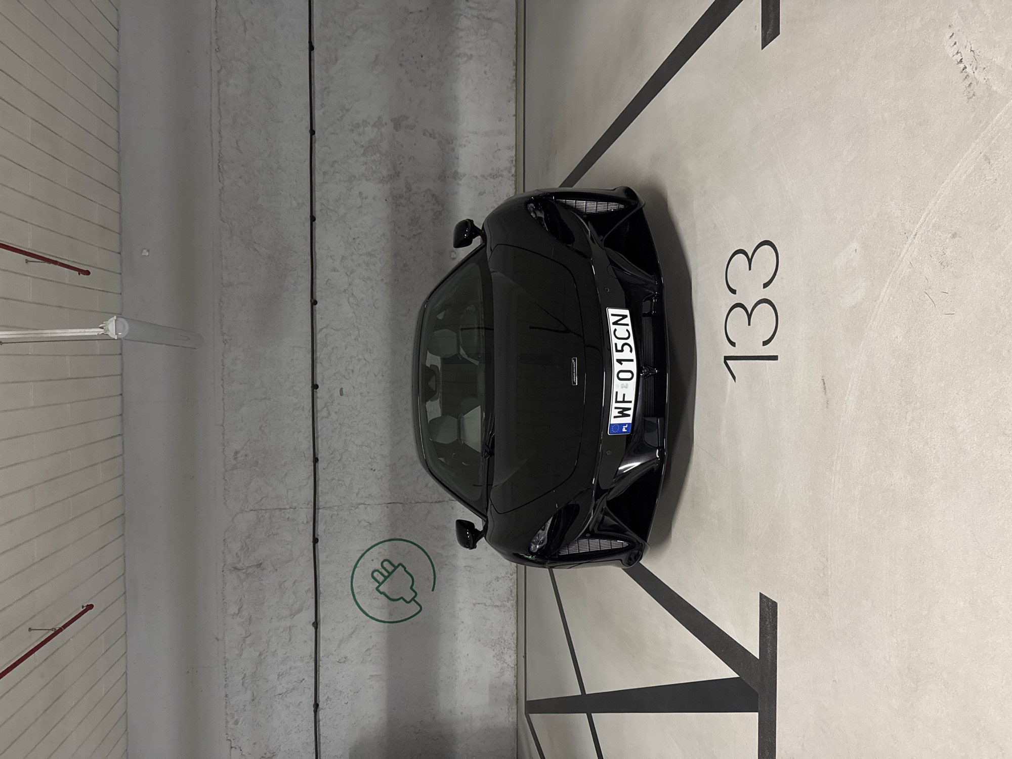The image shows a black car parked in a parking spot marked as number 133, with a green electric charging symbol, indicating it is an electric vehicle parking space. The license plate on the car is "WF 015CN" indicating that it is from Warsaw, Poland. The parking spot is in an indoor parking garage with concrete walls and floor.