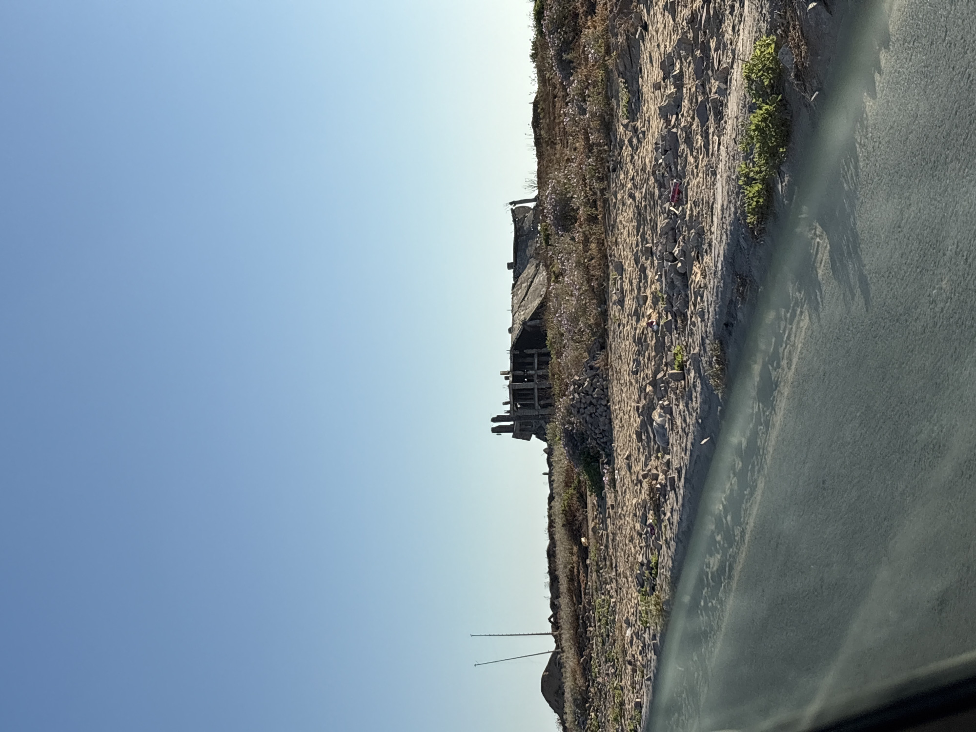The image shows a landscape with a clear blue sky and a rough, rocky terrain on the right side. There are remnants of a structure, possibly a building in disrepair, with exposed wooden beams and stone foundations. Vegetation, including small plants and flowers, grows sporadically on the rocky slope.
The perspective suggests the photo was taken from a vehicle, with a blurry, dark surface in the foreground, likely a car window or dashboard. The image is oriented vertically, making the sky appear on the left and the ground on the right. Two thin poles or antennas are visible in the distance against the sky.
The time of day appears to be daytime, with bright sunlight casting shadows. The weather seems clear and possibly warm. The overall scene evokes a sense of abandonment and decay, with the natural elements beginning to reclaim the man-made structure. No people are visible in the image. There is no visible text.