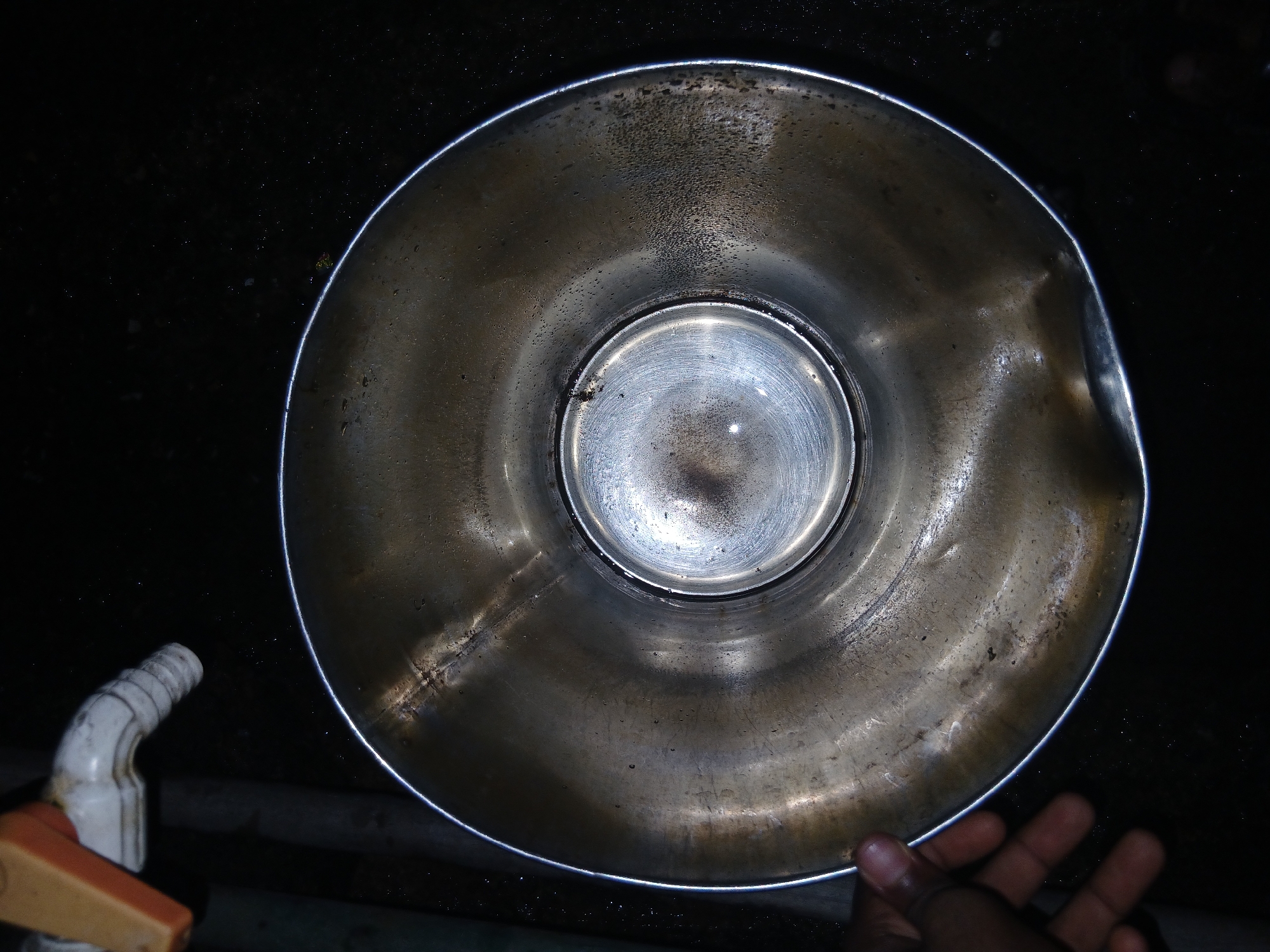 A top-down, close-up photograph shows a metallic, cylindrical container with water inside. The container appears to be stained and has a slight dent on the right edge. Light is reflected on the bottom of the container. The surrounding environment is dark. A tap with an orange handle is visible in the lower left corner. A hand, partially visible, is in the lower right corner.
There is no visual information present to determine the location within Lagos, Nigeria.