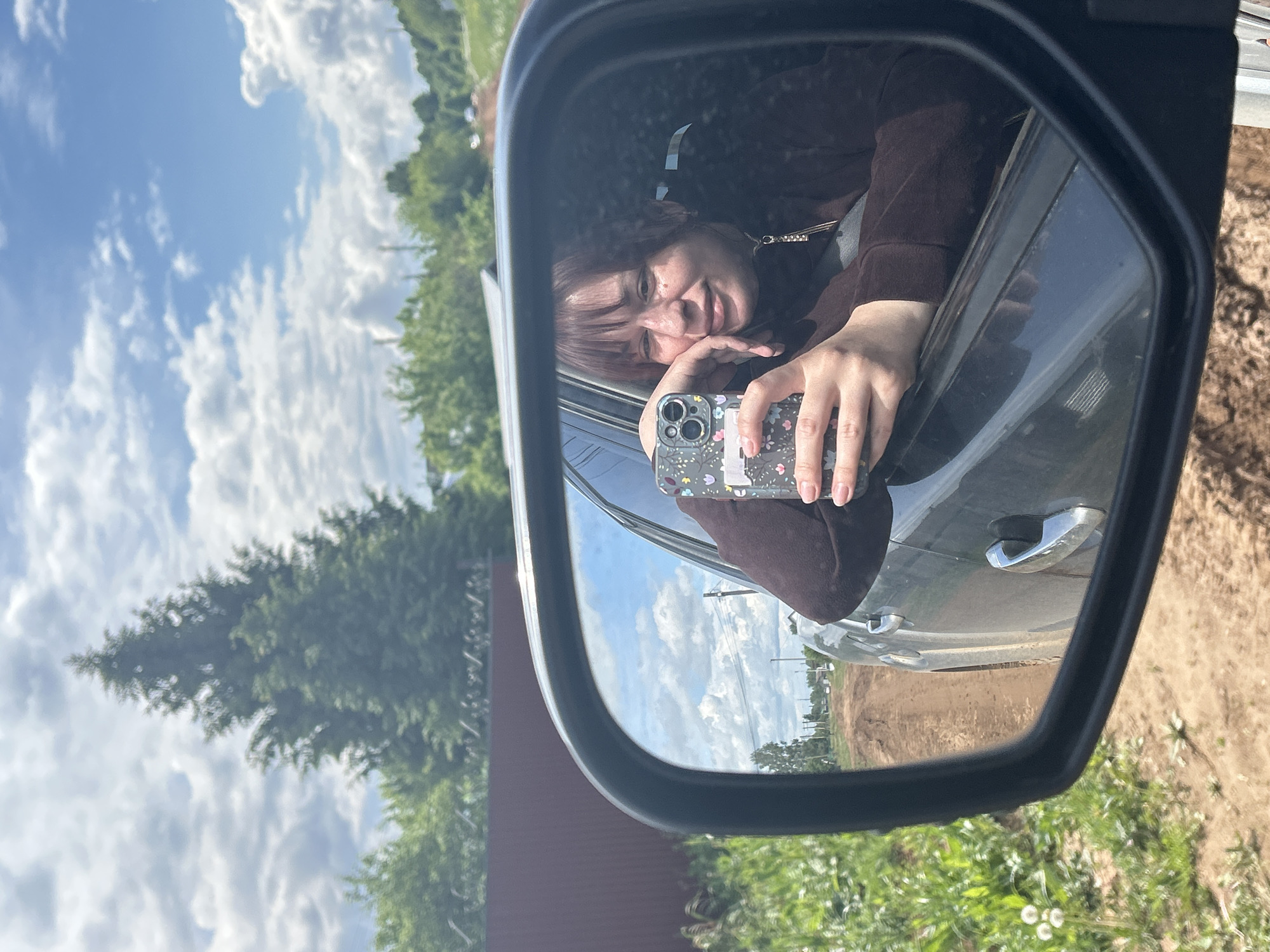 A rearview mirror shows a person in a brown top holding a phone with a patterned case, reflecting a view of the sky, trees, and potentially a field or cleared land. The person is smiling, and the mirror is attached to a car. The image captures a scene in Фоки, Russia, where the landscape includes vegetation and signs of construction or natural terrain.