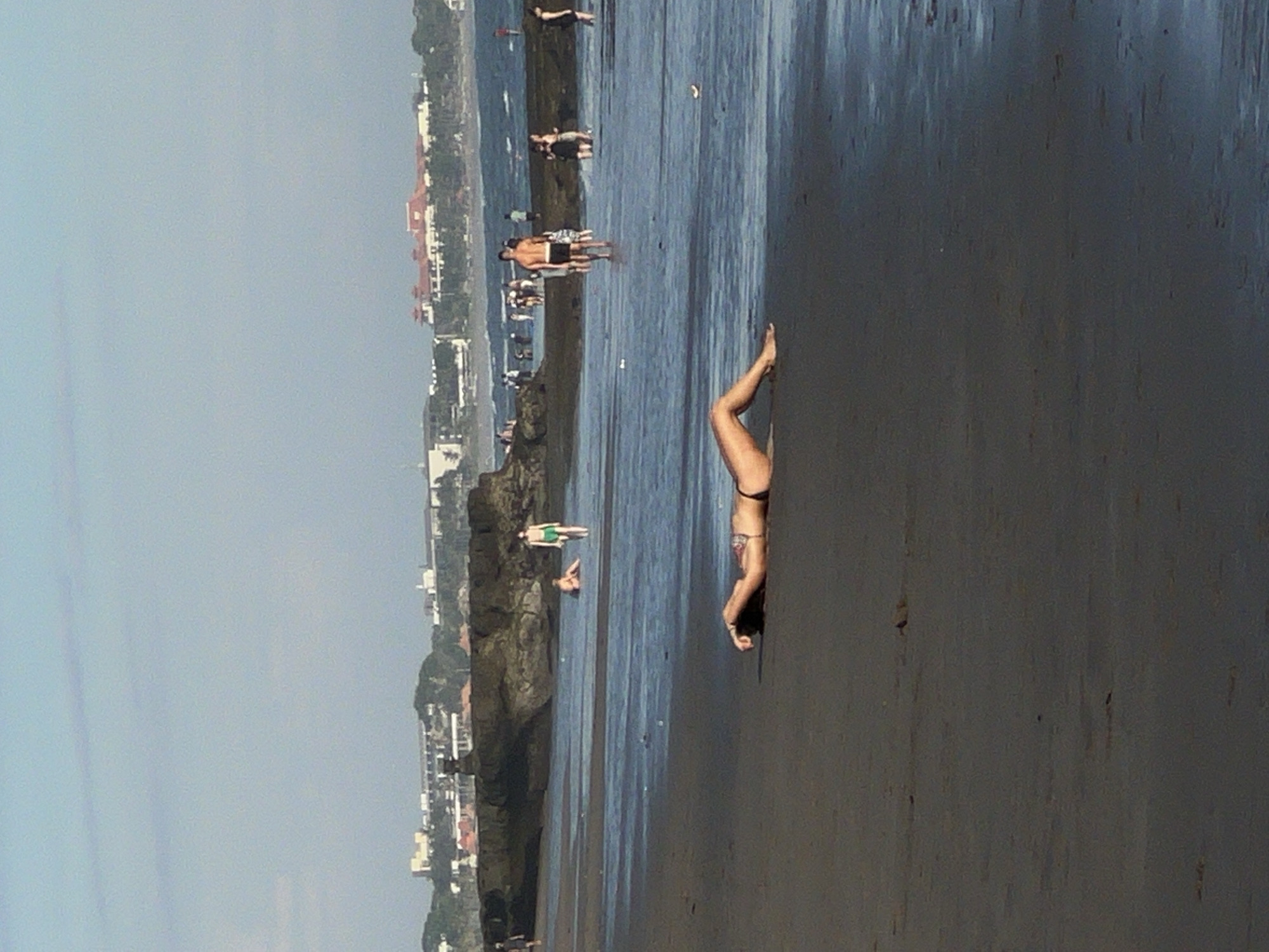 A woman in a bikini is doing a handstand on a beach. The beach is wet and dark, suggesting it's low tide. Other people are visible in the background, some standing near a rocky outcrop and others further down the beach. Buildings line the coastline in the background, characteristic of a populated area. The location is North Kuta, Indonesia.