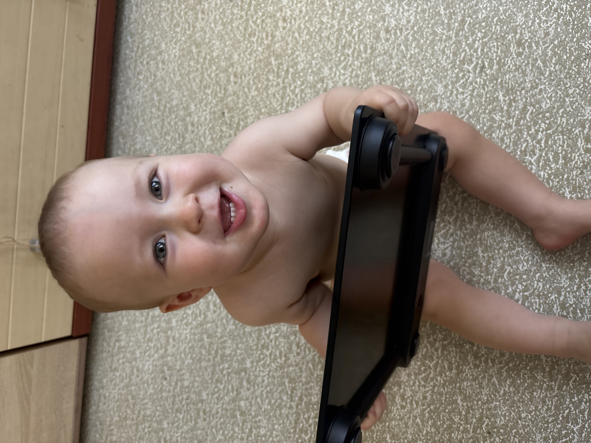 A baby is sitting on a carpeted floor and holding a black tablet computer. The baby is wearing a diaper and has light-colored skin and hair. The baby is looking at the camera and smiling, with teeth visible. The background consists of a wooden cabinet and the carpeted floor. The scene appears to be indoors, possibly in a residential setting. There is no information to indicate the location in the city of Naberezhnye Chelny, Russia.