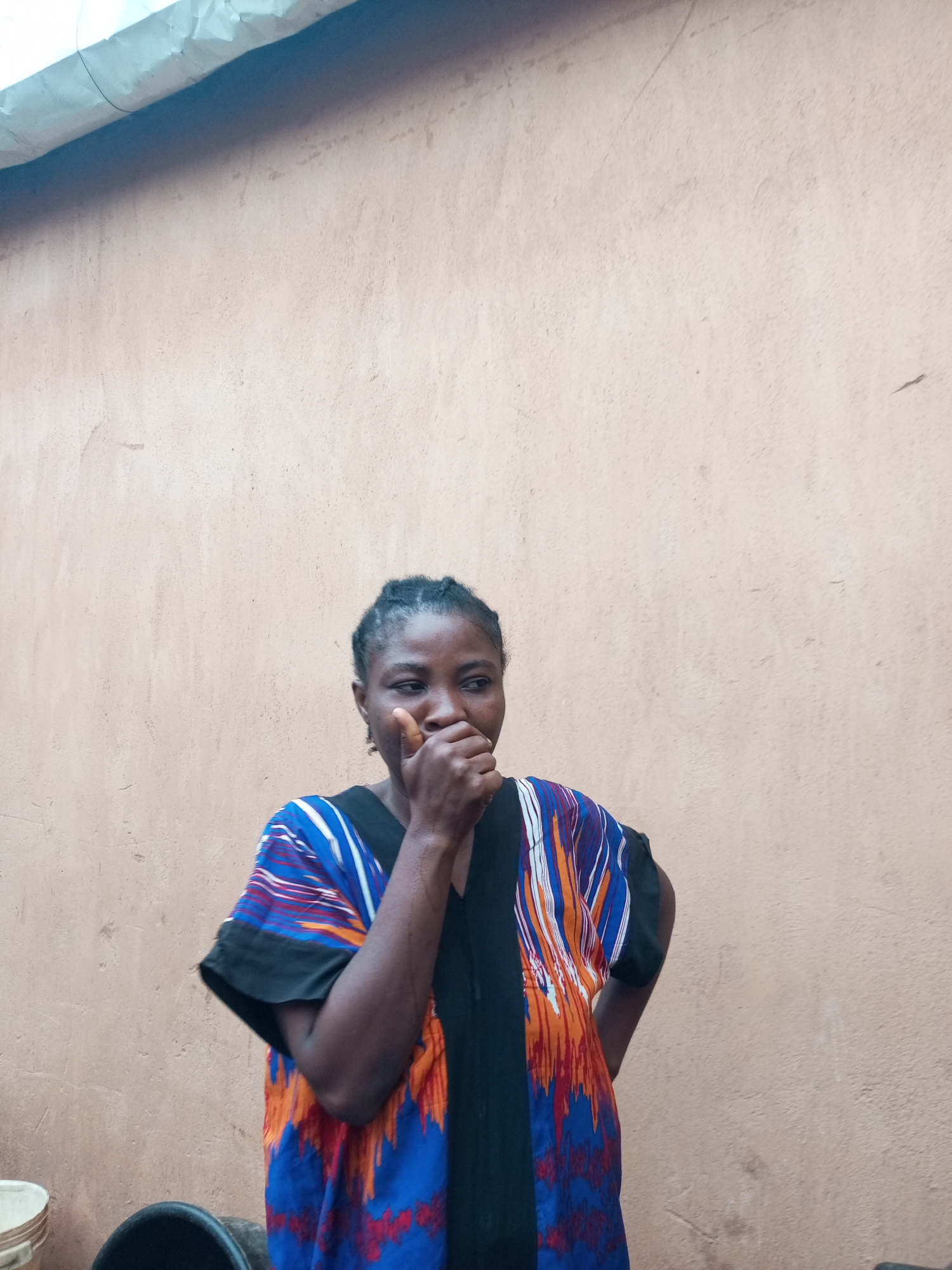 In Tempure, Nigeria, a dark-skinned woman stands outdoors against a tan, textured wall. She is wearing a colorful, patterned garment with black sleeves. Her right hand is raised, covering her mouth and nose, and her eyes are directed to her right. To the lower left of the frame, parts of two dark, circular objects are visible, possibly buckets or containers. The lighting suggests it is daytime.