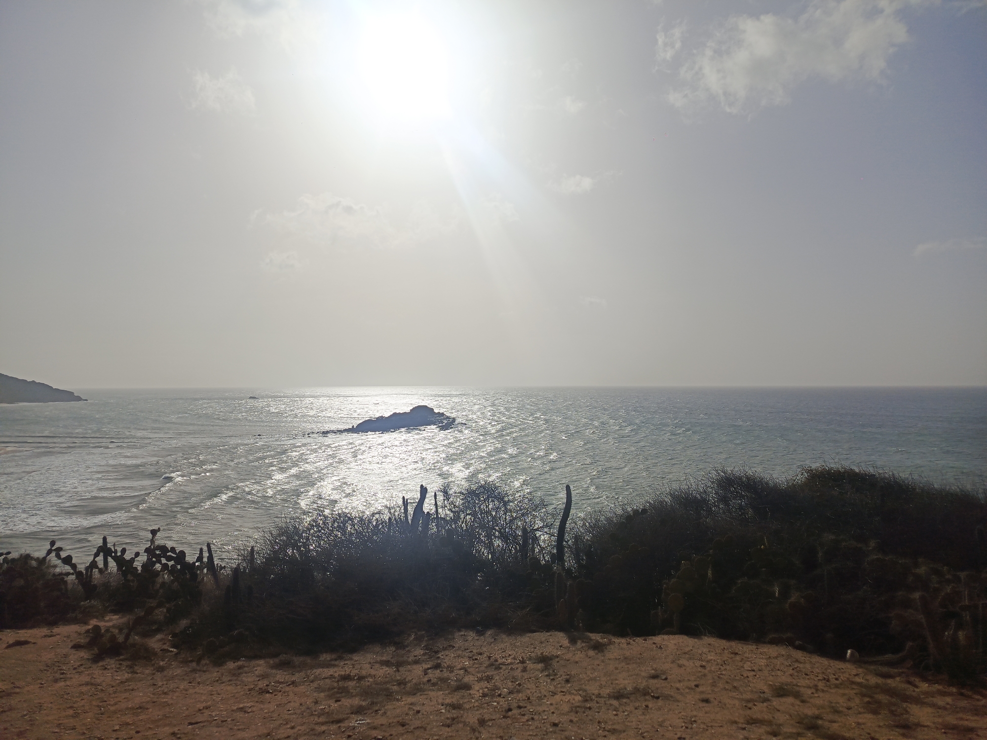 The image captures a coastal scene in Altagracia, Venezuela. The foreground features a sandy, reddish-brown patch of earth with scrubby vegetation, including cacti. The middle ground showcases the ocean, with sunlight reflecting on the water's surface, creating a shimmering effect. A rocky islet is visible in the distance. The horizon is somewhat obscured by haze, and the sky is bright with the sun. The overall atmosphere is sunny and dry.