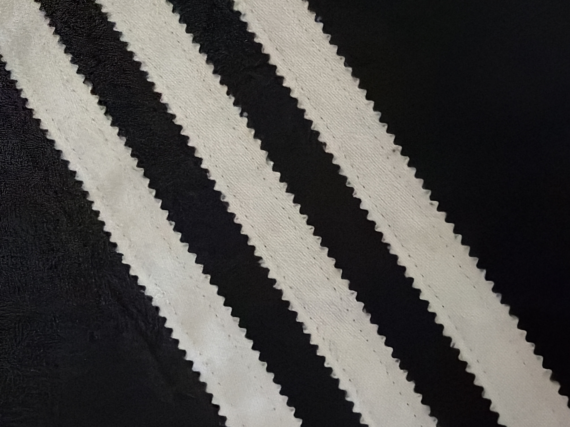 This is a close-up, abstract shot of a black fabric with three white, serrated stripes running diagonally across it. The stripes appear to be made of a textured fabric, possibly a towel or jersey material, with visible stitching along their edges. The black background is a smooth, matte material, possibly leather or a similar synthetic. The lighting is even, with no shadows indicating a specific time of day or location. The image focuses entirely on the texture and pattern of the fabric, creating a minimalist and graphic composition. There is no visible text in the image. The overall impression is one of simple, clean design and a focus on material contrast.