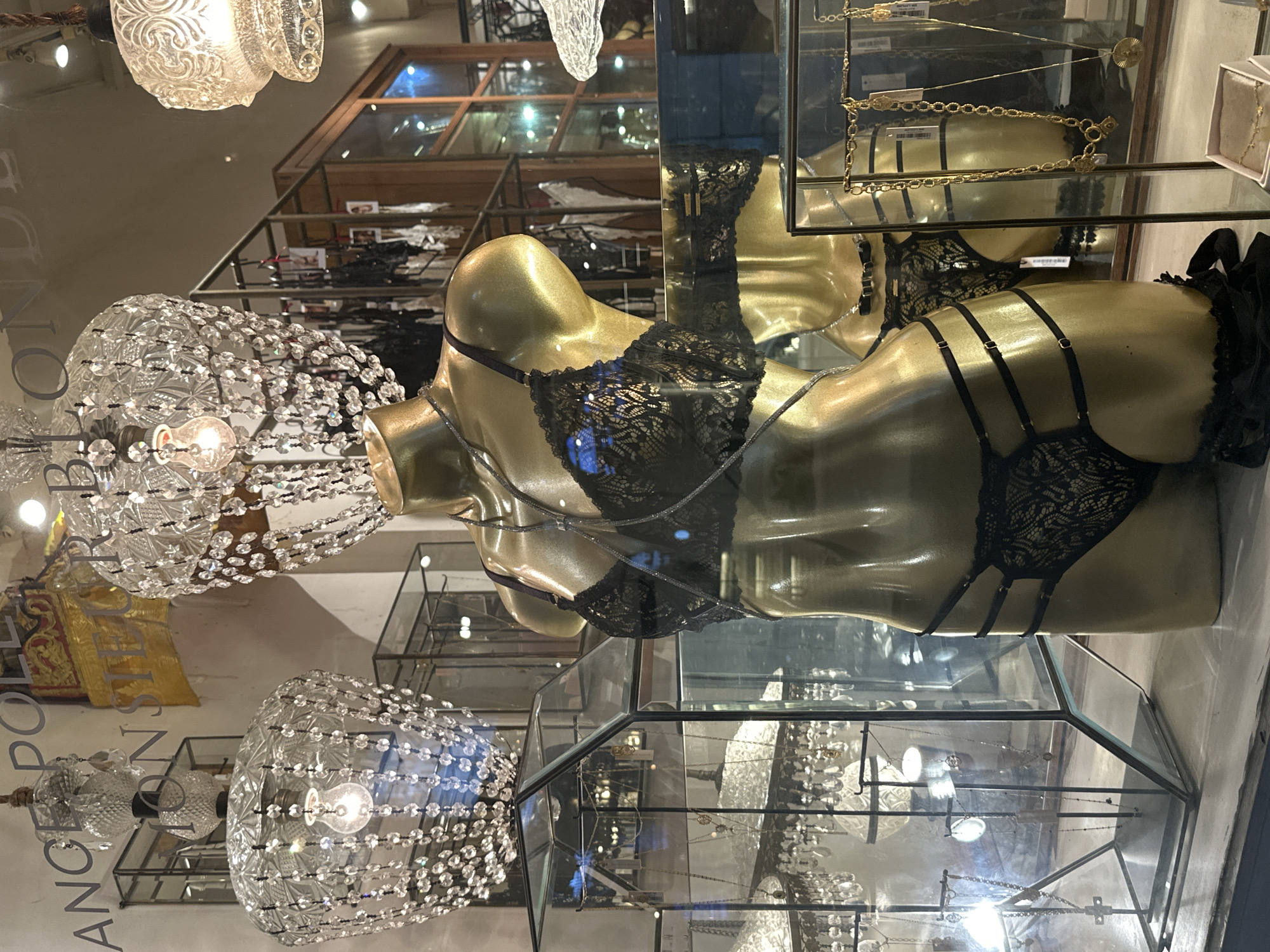 The image depicts a store window display in Kuta, Indonesia, featuring mannequins adorned with lingerie and jewelry. A golden mannequin, positioned centrally, wears a black lace bra and matching underwear with strappy details. It is accessorized with a delicate silver-toned necklace. Behind it, another golden mannequin is partially visible, also wearing lingerie.
The display is illuminated by decorative crystal chandeliers hanging from the ceiling. In the background, shelves are stocked with clothing, likely more lingerie, and other items. To the left, a glass display case showcases several necklaces, some with pendants, arranged on stands. The overall ambiance suggests a boutique specializing in intimate apparel and accessories. The store name "ANGELE POLLE MONSIEUR BLONDE" is visible on the window, indicating the store's identity. The scene is indoors, likely in a retail environment within Kuta.