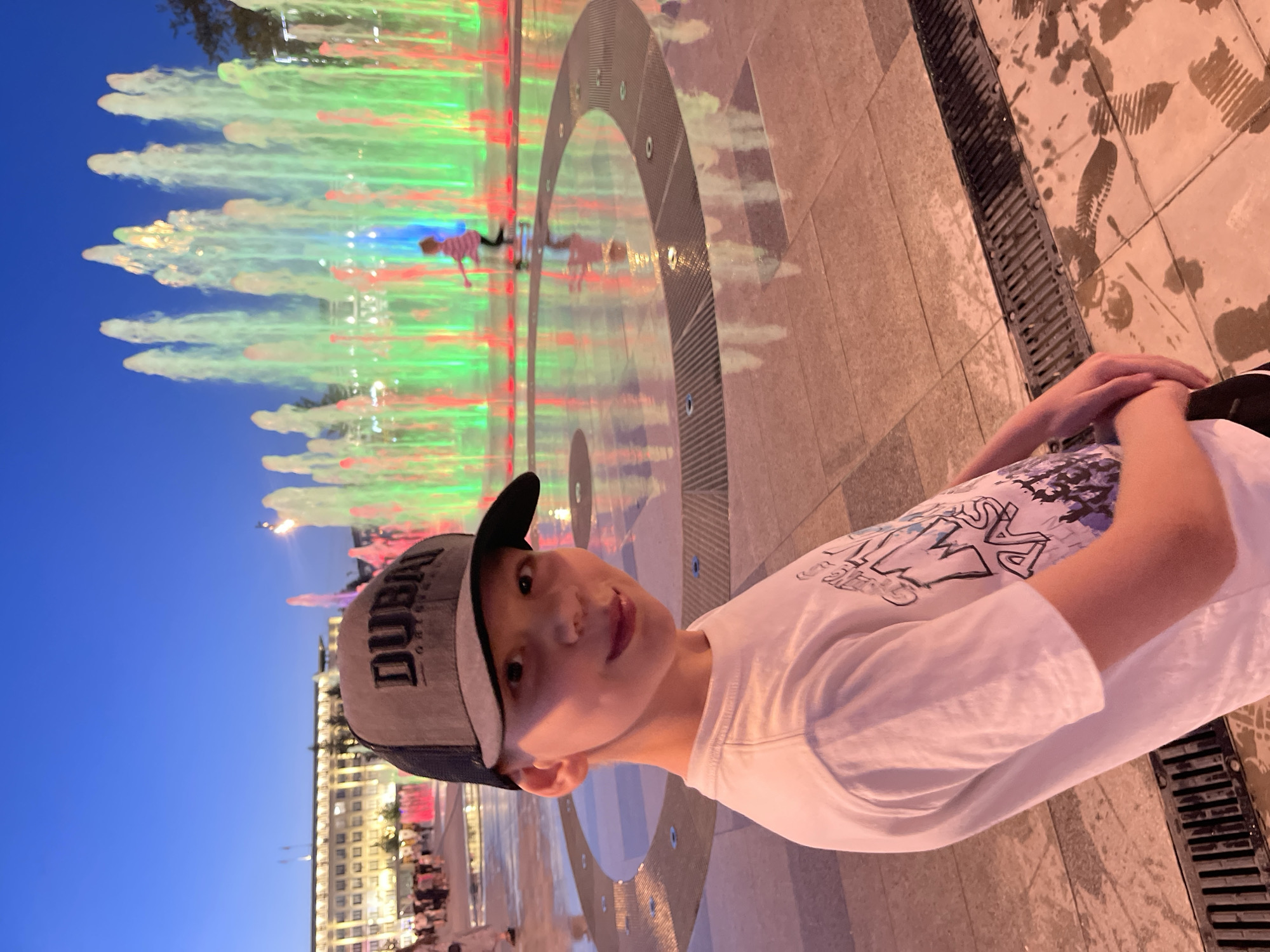 In this vertical, eye-level, outdoor, medium shot, a young boy wearing a white t-shirt and a gray and navy blue baseball cap with "DUBAI" written on it, looks towards the camera with a slight smile. He is positioned in the foreground, with his arms folded. Behind him, a vibrant water fountain displays a cascade of colorful lights, primarily green and red, illuminating the mist. The fountain is set against a backdrop of a clear, deep blue sky, indicating it is likely dusk or early evening.
The ground surrounding the fountain appears to be paved with light-colored tiles, some of which are wet, suggesting recent interaction with the water. A metal grate runs along the edge of the tiled area. In the distance, a large, illuminated building with many windows is visible, hinting at an urban or city setting. The overall atmosphere appears festive and recreational, as the illuminated fountain and the presence of other people in the background suggest a public gathering or a popular tourist spot. The lighting from the fountain and the building creates a warm, inviting ambiance.