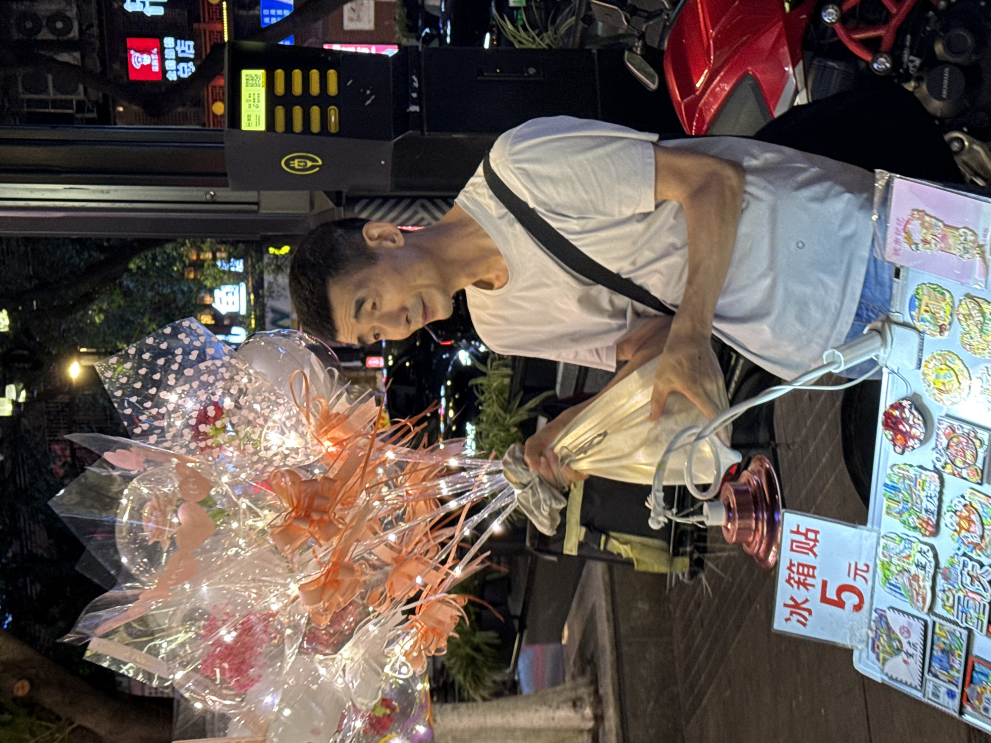A man stands on a city street in Yuzhong District, China, at night, holding a bouquet of illuminated balloons and a bag of merchandise. He is wearing a light-colored t-shirt and jeans, with a black strap across his chest. Behind him, a red motorcycle is parked. In the foreground, a stall displays various items, including stickers labeled "冰箱贴" (refrigerator stickers) priced at "5元" (5 yuan). The background features illuminated signs and building facades, indicative of an urban environment.