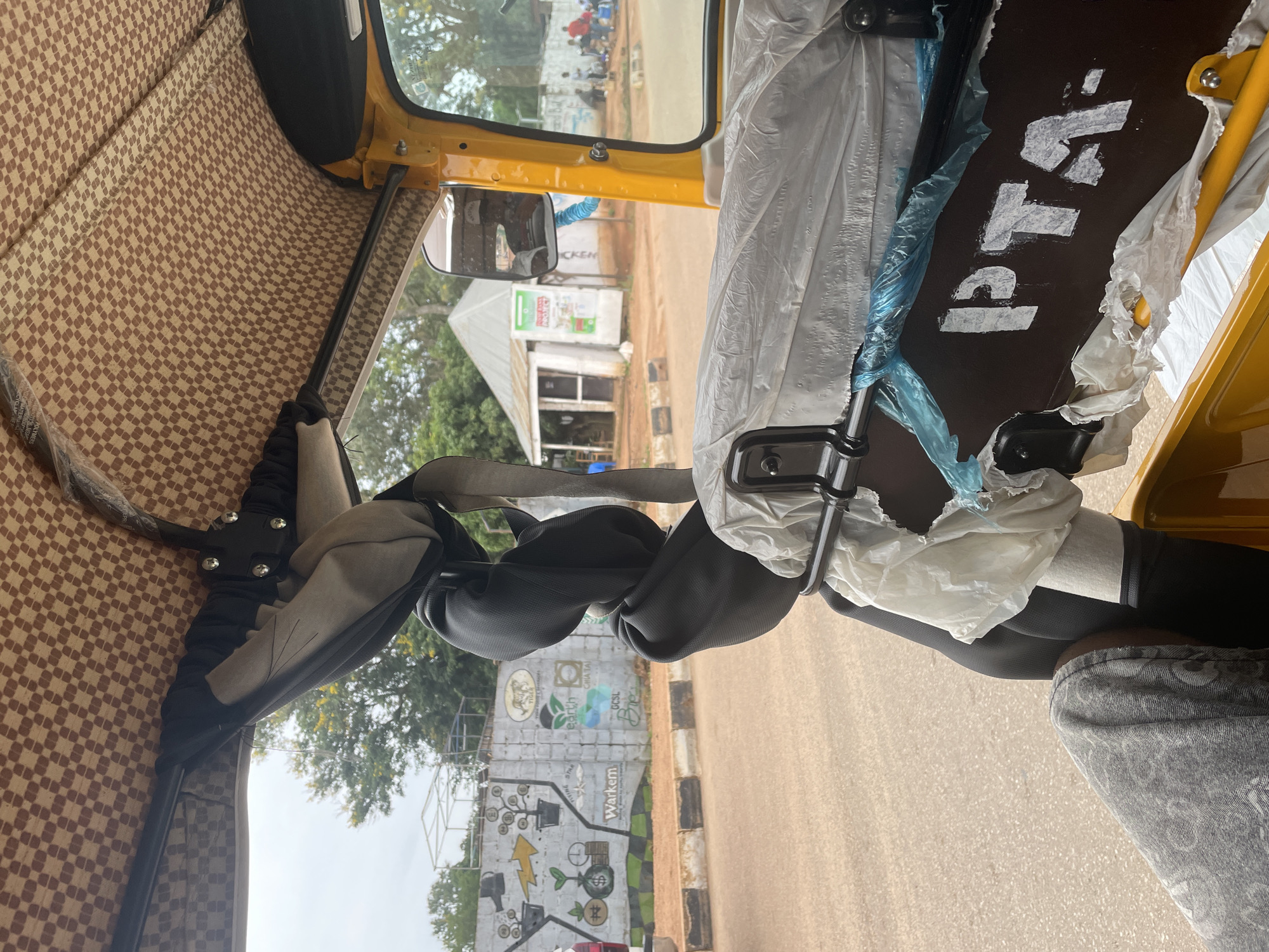 An interior view of a yellow auto rickshaw in Gura Top, Nigeria, shows a checkered brown fabric ceiling. A black padded bar runs across the frame, draped with a folded piece of black fabric. To the right, a dark brown panel with white lettering reads "PTA-". The panel is partially covered by white plastic sheeting and blue tape. Through the open side of the auto rickshaw, a grey paved road is visible, bordered by a dirt shoulder and a concrete wall decorated with murals depicting plants, coins, and logos such as "Warkem" and "Earth". Further in the background, a small white building with a grey roof stands near trees. Several people are visible near the building and along the wall, some sitting and others standing. The sky is overcast.