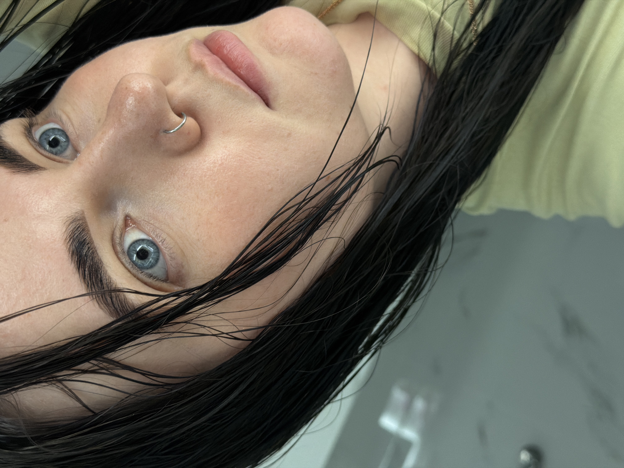 A close-up portrait of a woman's face, slightly off-center and rotated, showcasing a fair complexion, blue eyes, and dark, wet hair. The woman has a nose ring. She wears a light yellow top with a visible gold chain. The background shows a blurred white or light gray surface. The image provides no specific context to identify the location as Chaikovsky, Russia.