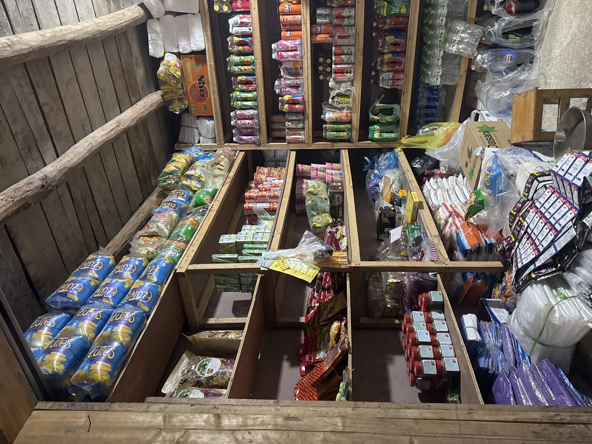 The image depicts the inside of a small store, possibly in the city of Lũng Cú Commune, Vietnam, judging by the products stocked and the rural setting. The store is in a wooden structure, with shelves built into the walls. The shelves are stocked with various goods, including packaged snacks (like "Lutes" brand chips), instant noodles ("Nho" brand), canned drinks (Coca-Cola, Fanta), bottled water ("Dasani"), condiments, plastic bags, and other household items. The arrangement is somewhat cluttered, with goods filling every available space. The lighting is dim and appears to be natural, coming from an unseen source. The overall impression is of a small, basic store serving a local community.