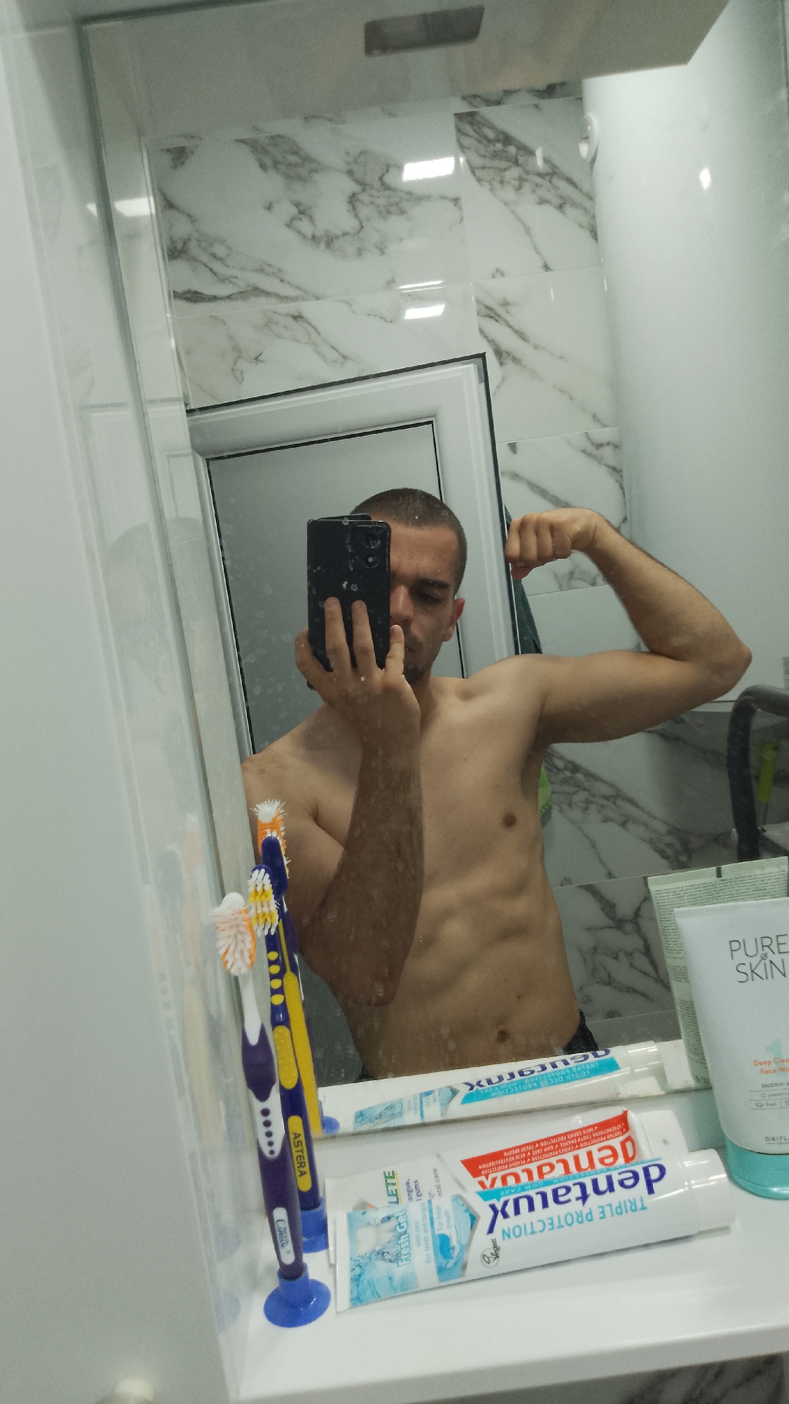 A man is taking a selfie in a bathroom mirror. He is shirtless and flexing his biceps. The bathroom has white marble-patterned walls. In the foreground, there are toothbrushes and tubes of toothpaste, including "dentalux TRIPLE PROTECTION" and "ASTERA." Also, a tube of "PURE SKIN" is visible. Based on the brands of toothpaste, the location could be Bulgaria. The city is unknown; however, the image does not contain definitive evidence of the city of Yambol.