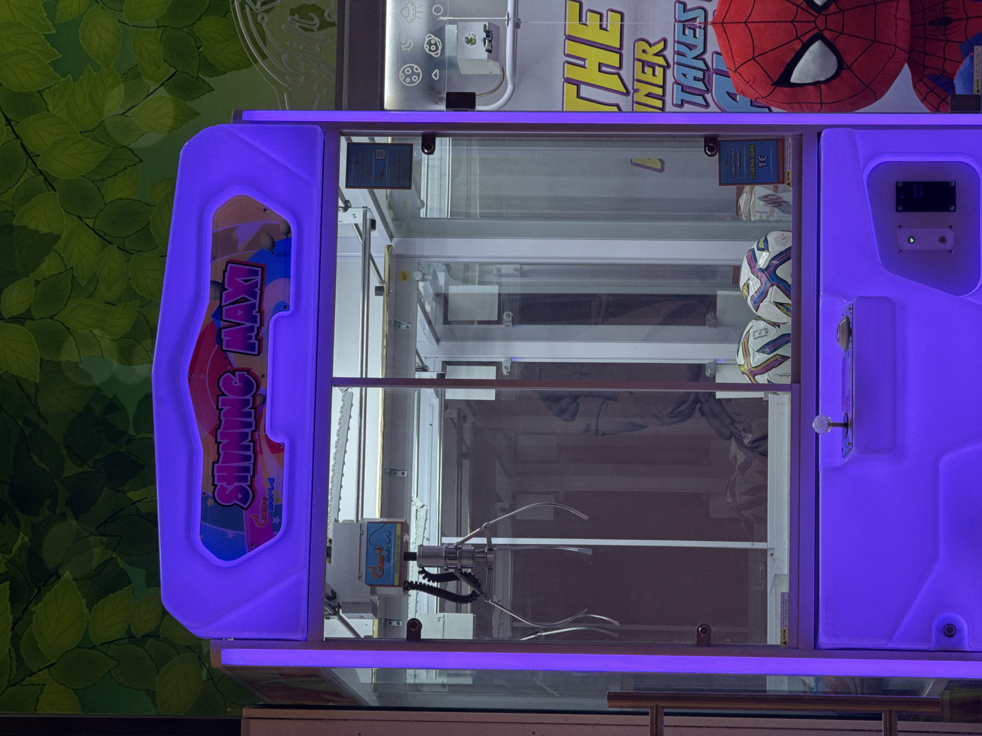 Here is a description of the image:
The image shows a "SHINING MAXI" claw machine, illuminated in purple light. Inside the machine, there are at least two soccer balls and a Spider-Man plush toy. A claw is visible, ready to grab a prize. The machine also has signage indicating the game's cost. The background includes a green leafy design and other elements suggesting this is located in an entertainment or arcade area.