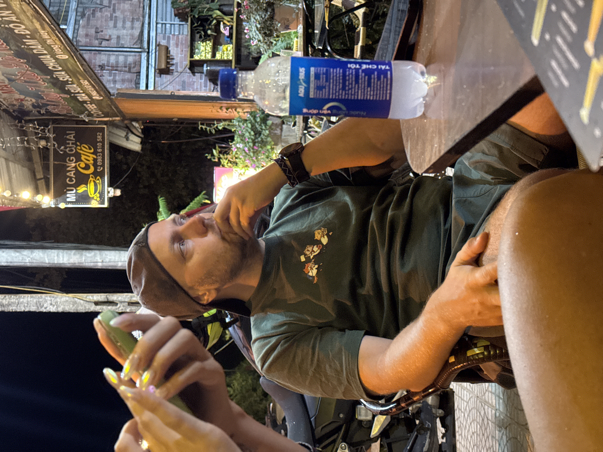 The image captures a casual nighttime scene at an outdoor café in Mù Cang Chải, Vietnam. A man, wearing a dark green t-shirt with a small graphic, a dark cap, and a watch on his left wrist, is seated and looks contemplatively towards the left. He has a beard and appears relaxed. Adjacent to him on a dark table surface is a plastic Aquarius water bottle, featuring Vietnamese text including "TẢI CHẾ TÔI" and "100 g vận động."
In the foreground, the hands of another person, adorned with long, light-colored nails, are holding a light green object, possibly a vape device or a phone. A partially visible leg or thigh of this individual is also seen on the right side of the frame.
The setting is dimly lit by artificial string lights and overhead fixtures, indicating it is nighttime. Greenery and plants are visible in the background against a brick wall, creating a natural and inviting atmosphere. Parts of what appears to be a scooter or motorcycle are visible beneath the table.
A prominent black sign on the left clearly identifies the establishment as "MÙ CANG CHẢI Café," also listing services like "COFFEE - TRÀ SỮA - SỮA CHUA DẦM - ĐỒ ĂN" (Coffee - Milk Tea - Yogurt with Toppings - Food), along with a phone number "0983 610 582" and the name "Vàng Thị May." The overall scene portrays a relaxed evening gathering at a local Vietnamese café.