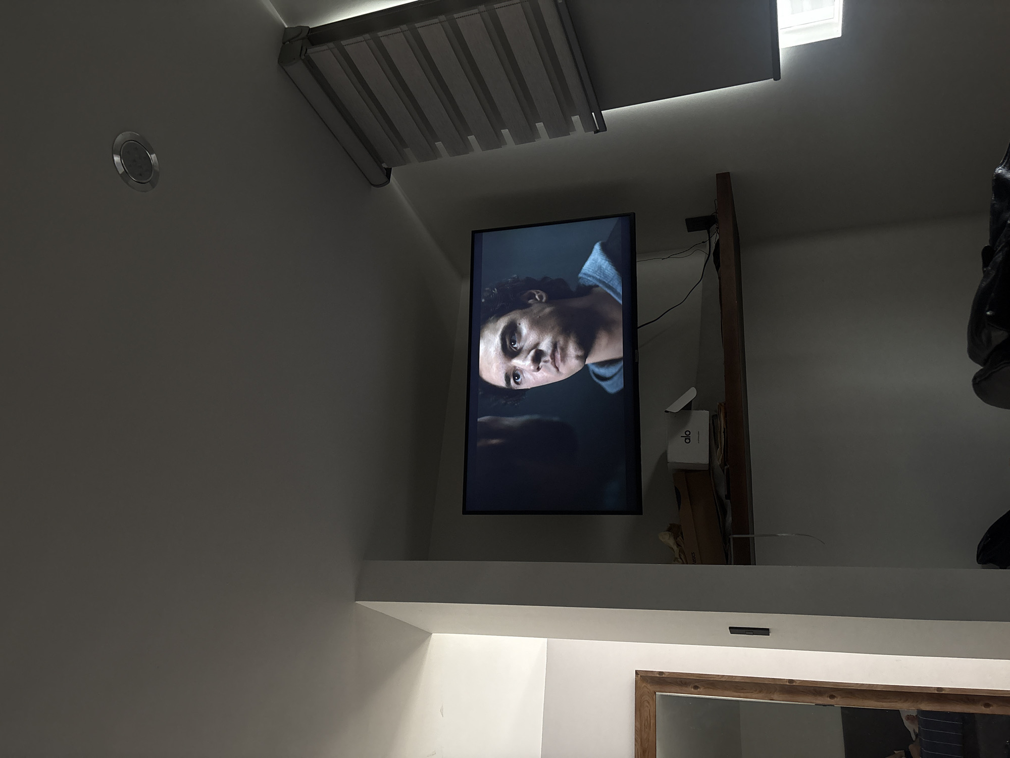 The image shows a dimly lit interior space in San Bernardino Tlaxcalancingo, Mexico. A television screen displays a close-up of a person's face. The TV is mounted on a wall above a shelf. Above the TV are ceiling fixtures and what appears to be a window with blinds. A mirror and other miscellaneous objects sit on the shelf.