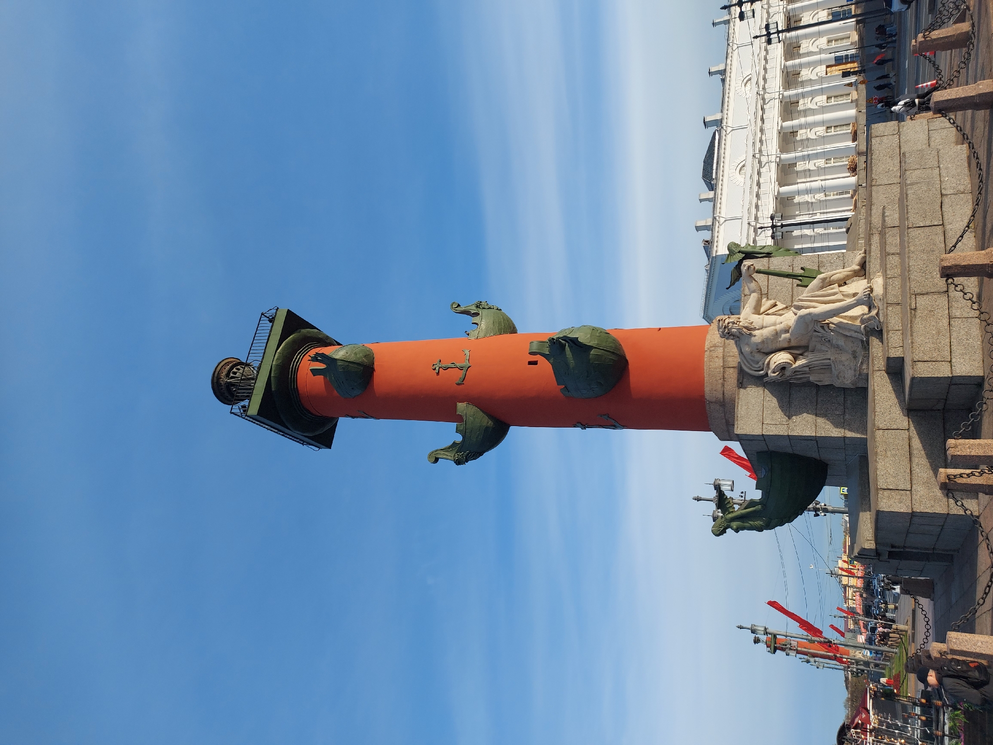 The image shows the Rostral Columns in Saint Petersburg, Russia, against a blue sky with faint clouds. The red columns are adorned with bronze ship prows. The base features sculptural groupings. A white building with columns is partially visible in the background, along with street-level views of people, vehicles, and other structures.