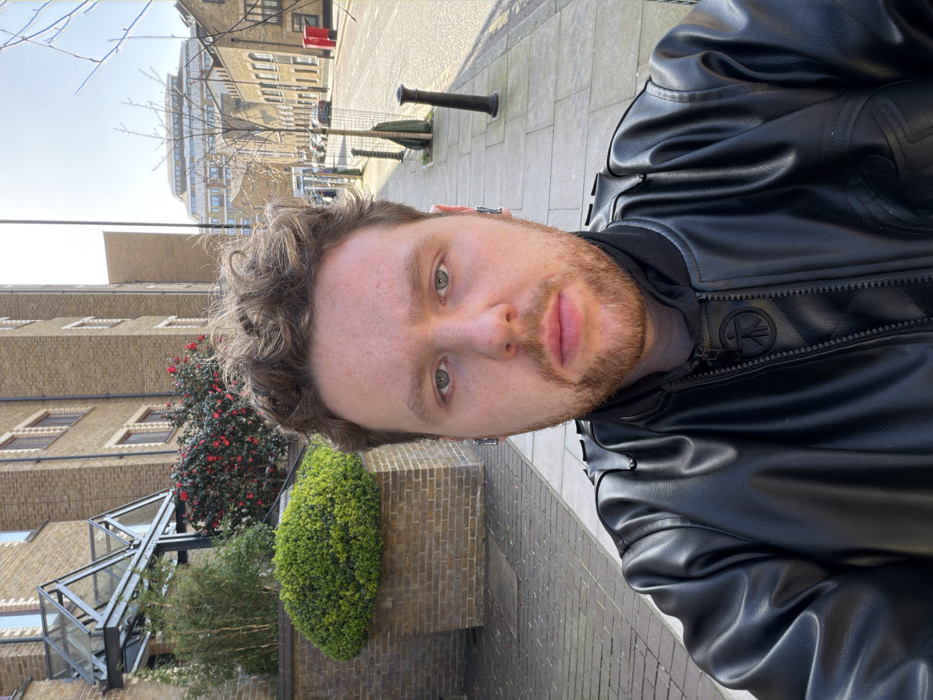 A male individual is depicted in a close-up selfie, positioned centrally and occupying the lower two-thirds of the frame. He has light skin, medium-length curly brown hair, a short beard, and light-colored eyes, looking directly at the camera with a neutral expression. He wears a black leather bomber jacket over a dark garment with a raised collar; a circular emblem with a stylized figure is visible on the jacket's zipper pull. Silver-colored wireless earbuds are present in both ears.
The scene is an urban street in London, United Kingdom, under clear daytime conditions with strong sunlight casting shadows. Behind the individual, a paved stone sidewalk extends into the background. To the immediate left, a multi-story brick building features a distinctive glass and metal canopy entrance, a large brick planter with a trimmed green bush, and a flowering shrub with red blossoms. Further down the street on the right, additional brick buildings line the road. Street furniture includes black bollards, bare deciduous trees protected by metal cages, and a red pillar postbox. Parked cars are partially visible along the street. In the far distance, a modern building with a multi-tiered roofline is visible against a bright, clear sky.
