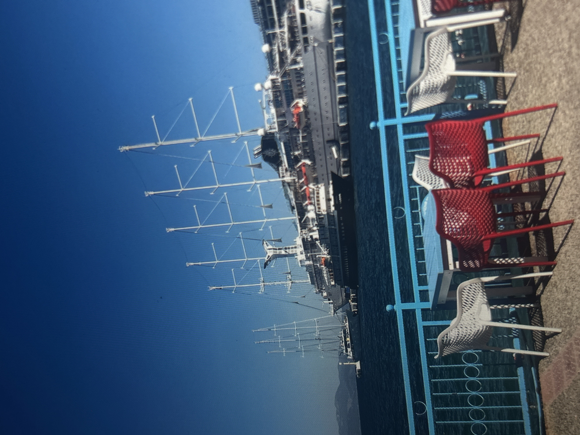 The image shows a waterfront scene with large sailing ships docked in the background, under a clear blue sky. In the foreground, there is an outdoor seating area with several tables and chairs, including white and red plastic chairs. A blue railing separates the seating area from the water. The location is identified as Yagodovo, Bulgaria, however, the visual evidence within the image does not support this specific location and instead appears to depict a coastal or harbor setting. The scene is tranquil and appears to be during daylight.
