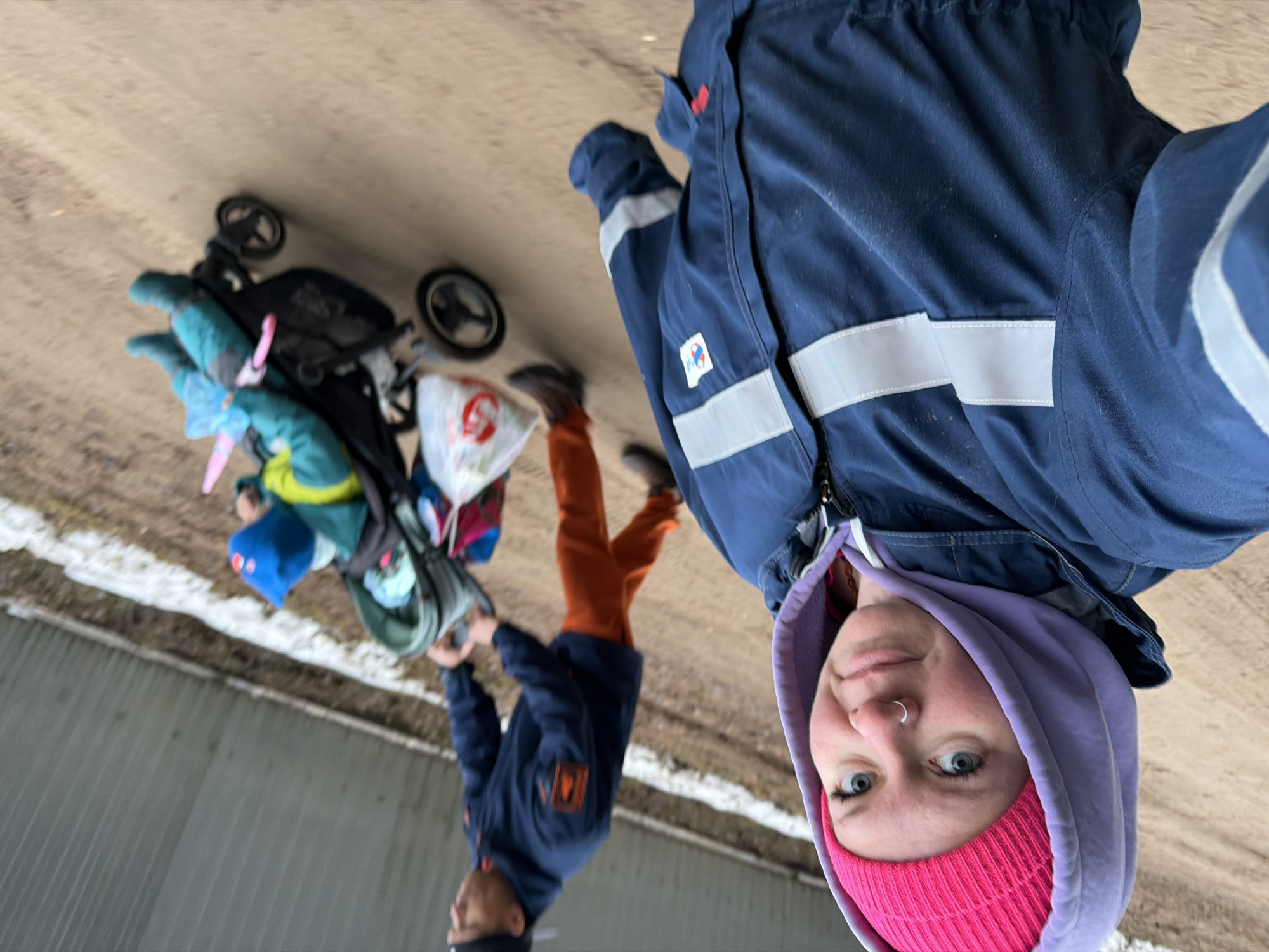 The image shows a selfie taken by a person wearing a pink beanie and a lavender hoodie under a blue jacket with reflective stripes. The person has blue eyes and a nose ring. Behind them, partially out of focus, is a man in a blue jacket and orange pants, interacting with a stroller carrying a child. The stroller is laden with bags and belongings. Patches of snow are visible on the ground. The location appears to be outdoors on a paved surface in Chaikovsky, Russia.