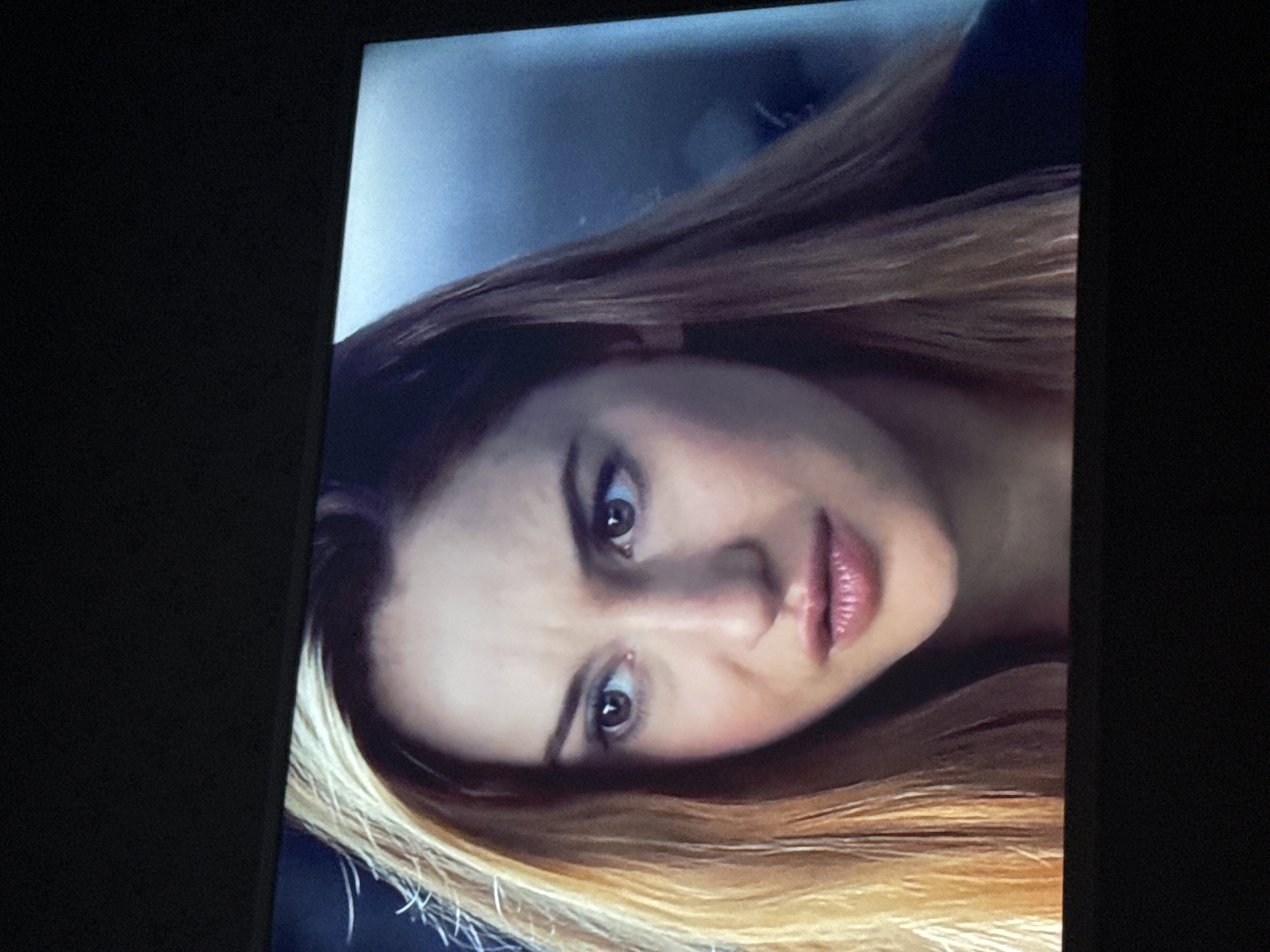 A medium close-up shot depicts a woman with fair skin and brown eyes looking directly at the camera with a slightly concerned expression. She has light brown hair with blonde highlights. The background is blurred and indistinct, but suggests an indoor setting. The image appears to be a photograph of a television screen. The location is not discernible from the image.